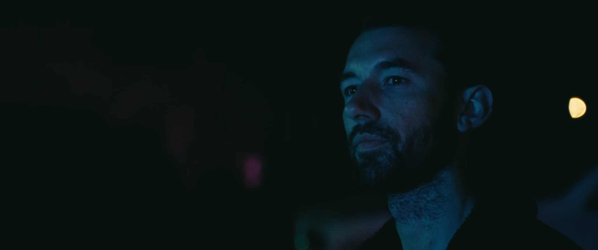 A man with a beard looks to the side in a dark setting with minimal lighting.