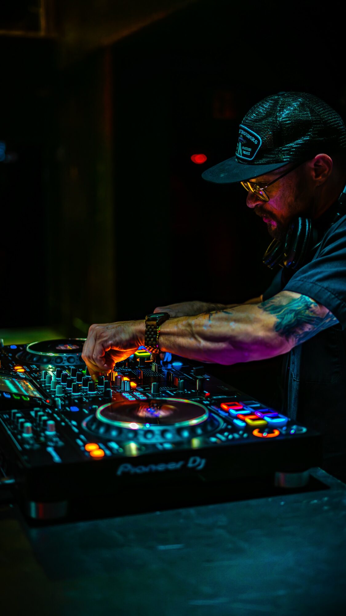 DJ wearing a cap and glasses, operating a DJ controller in a dark setting with colorful lights.