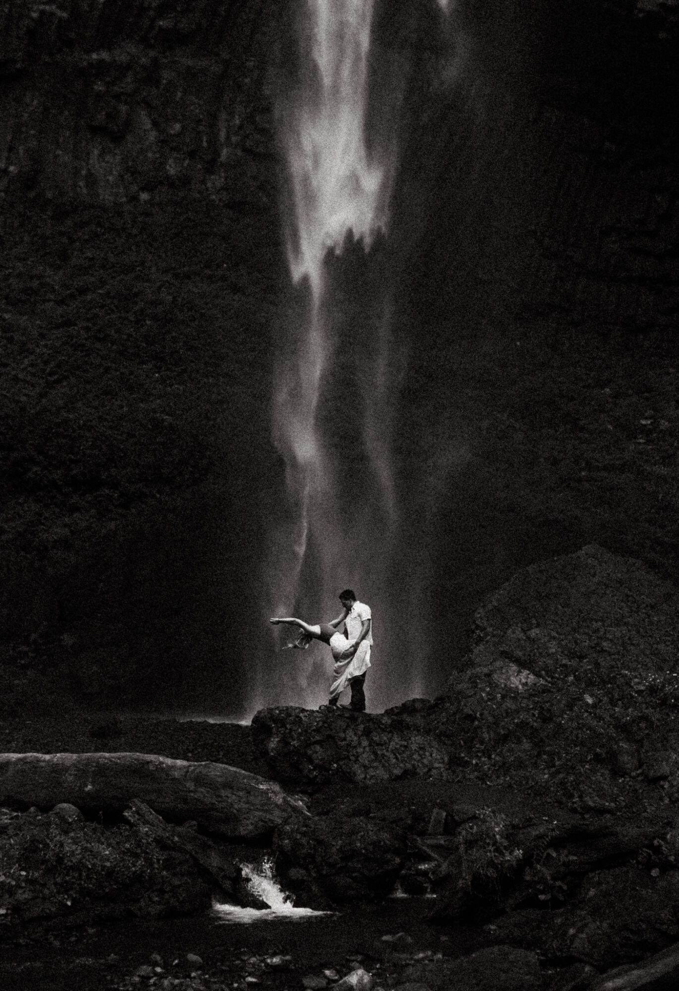 Person standing near a waterfall with water falling from above, surrounded by dark rocks.