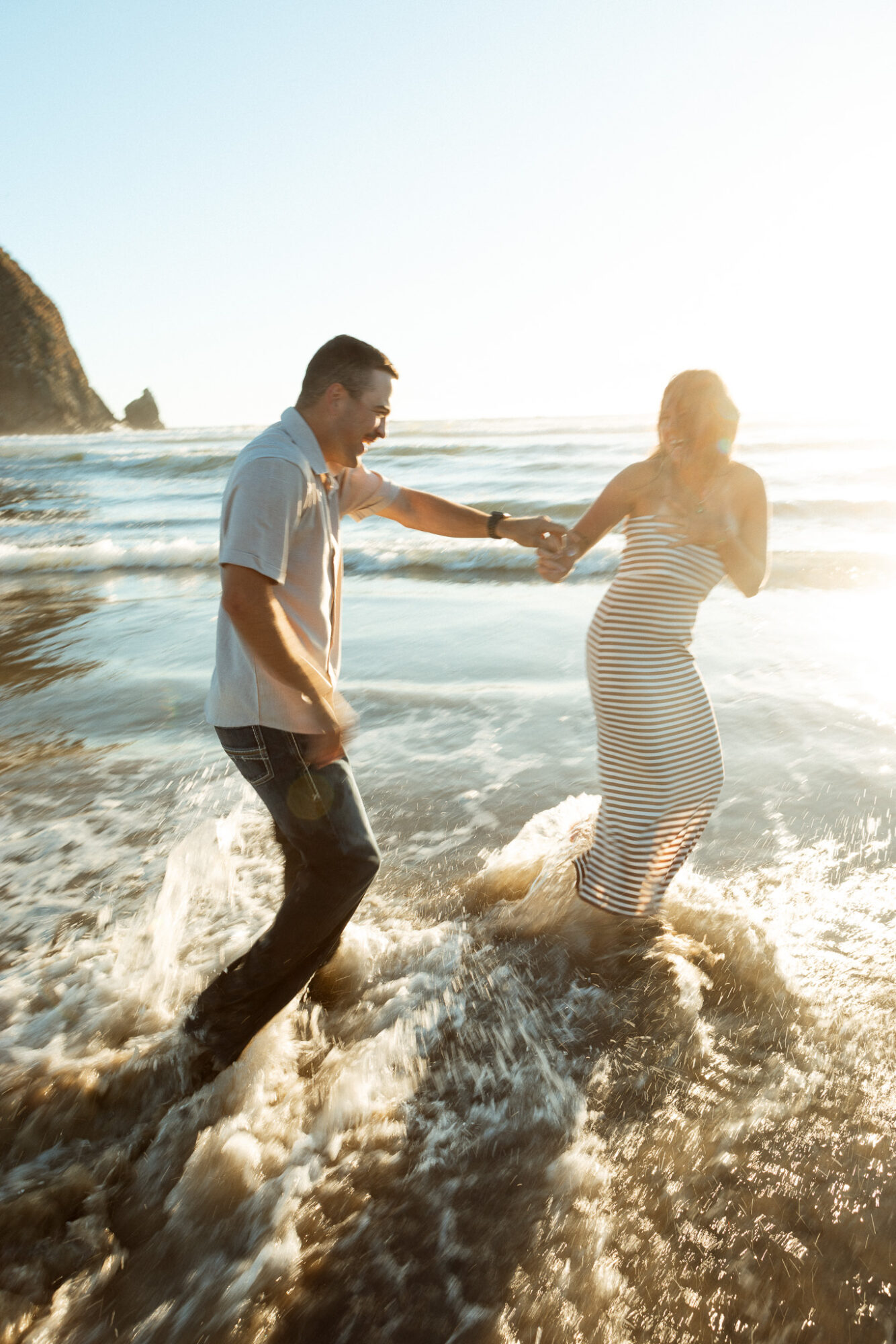 A man and woman holding hands in shallow ocean water at the beach, with a rocky cliff in the background, sunlight shining brightly.