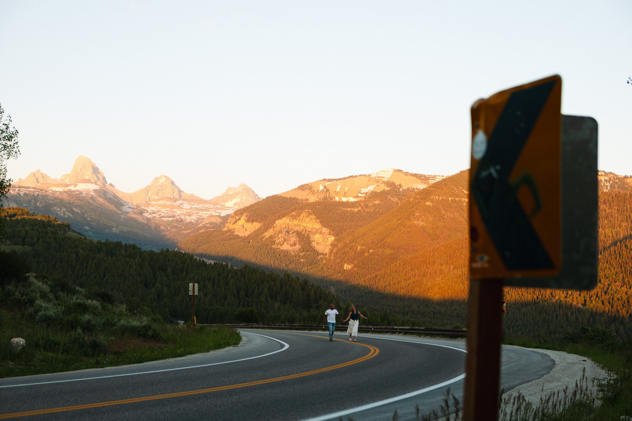 Curving mountain road with a warning sign, two people walking, mountains in the background, sunset lighting.