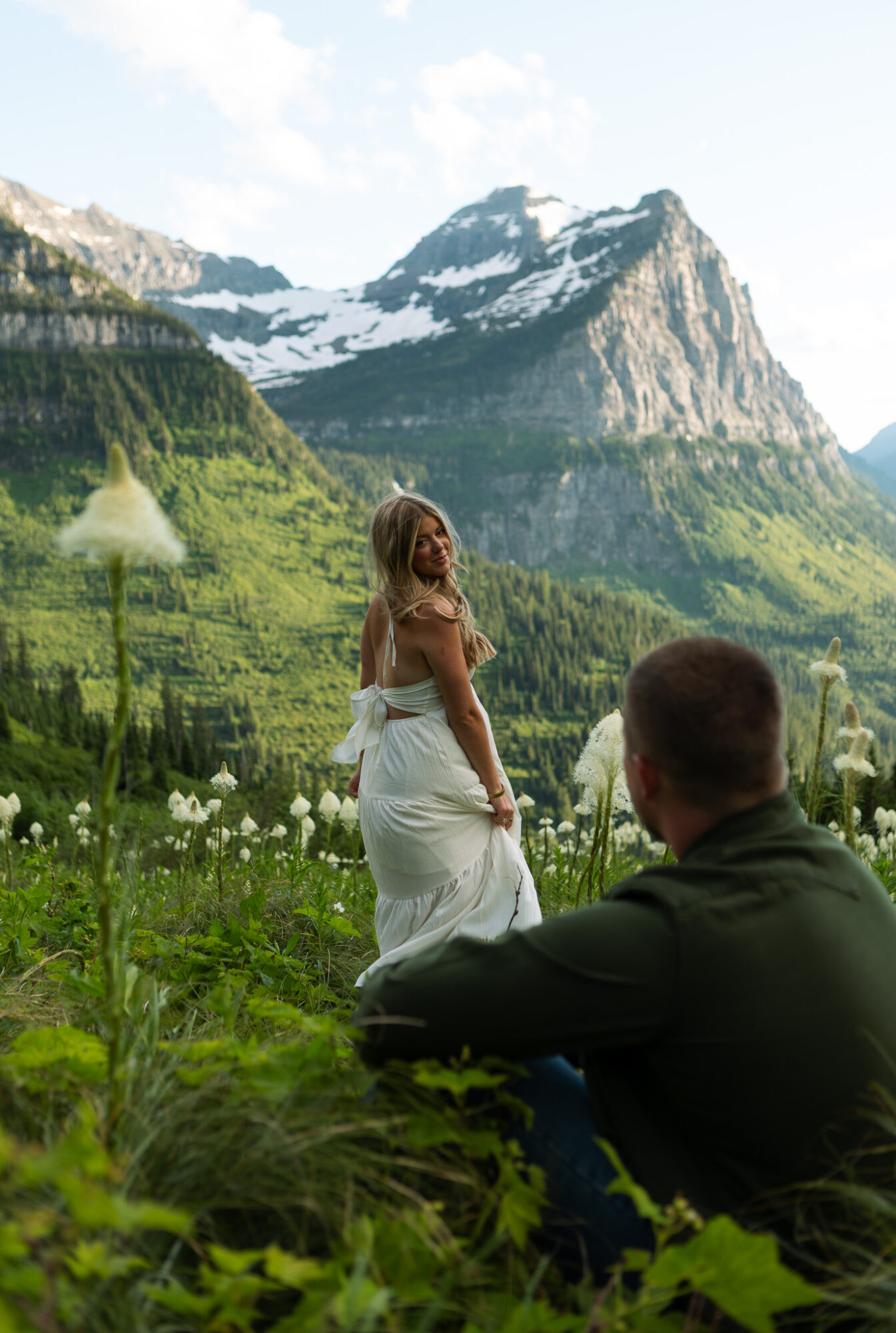A woman in a white dress stands in a green field with mountains and snow in the background, while a man sits nearby.