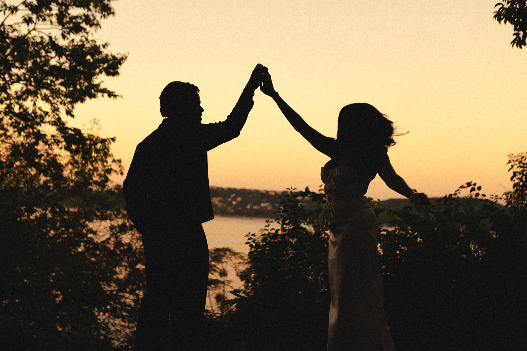 Silhouettes of two people high-fiving outdoors at sunset near a body of water with trees in the background.