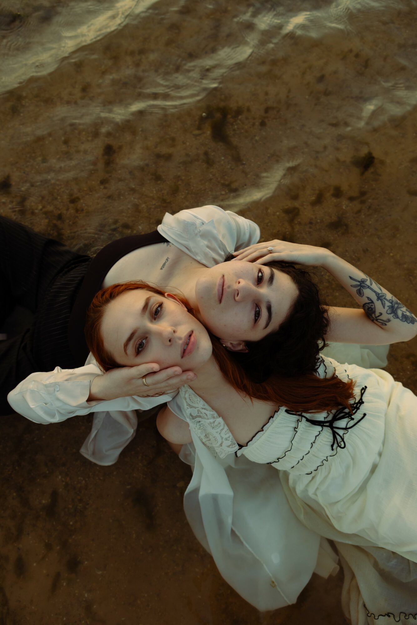 Two women lying on the ground, one resting her head on the other's shoulder, with a rocky background.