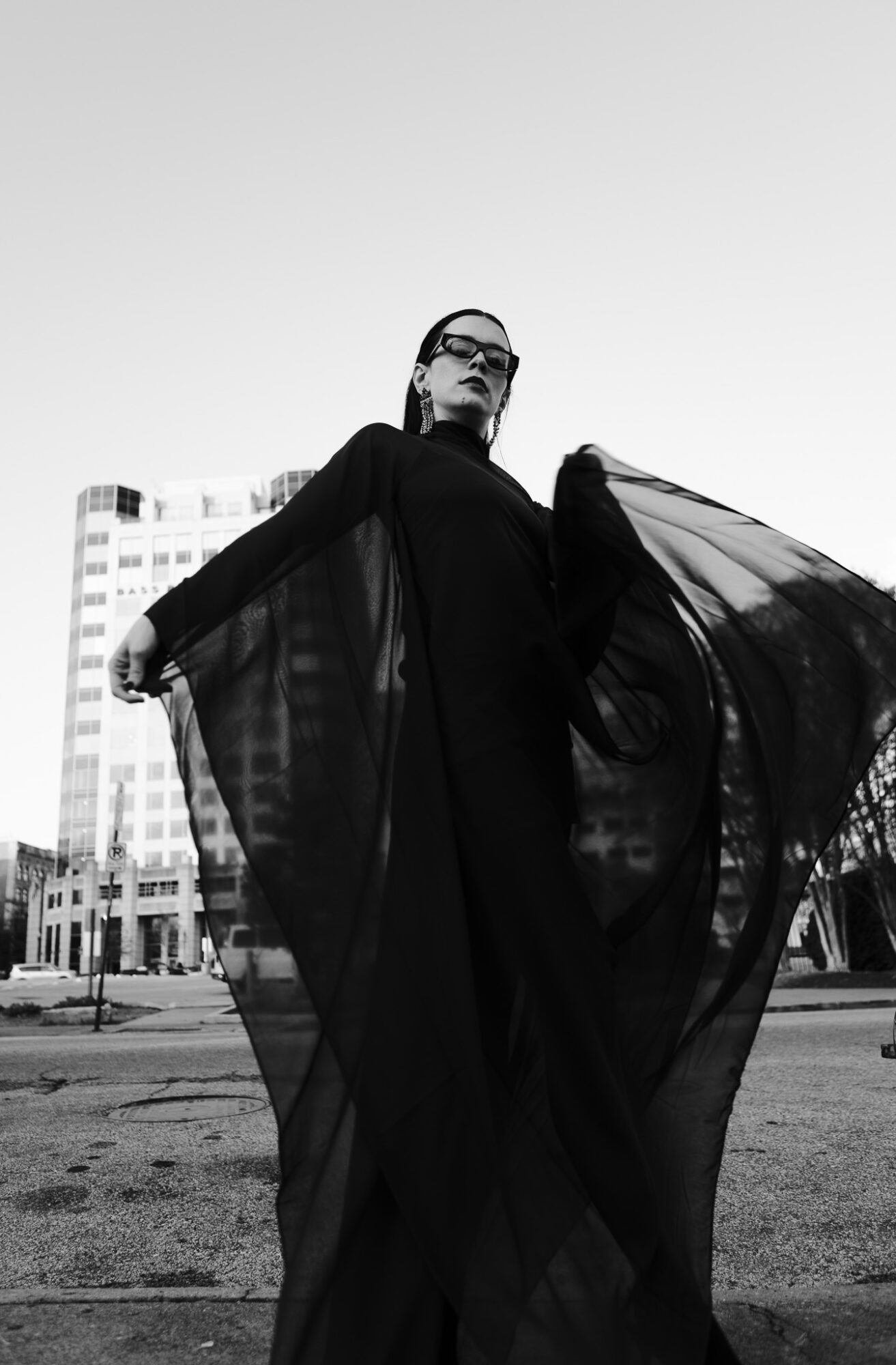 Woman in black dress holding flowing fabric outdoors with city buildings in background.