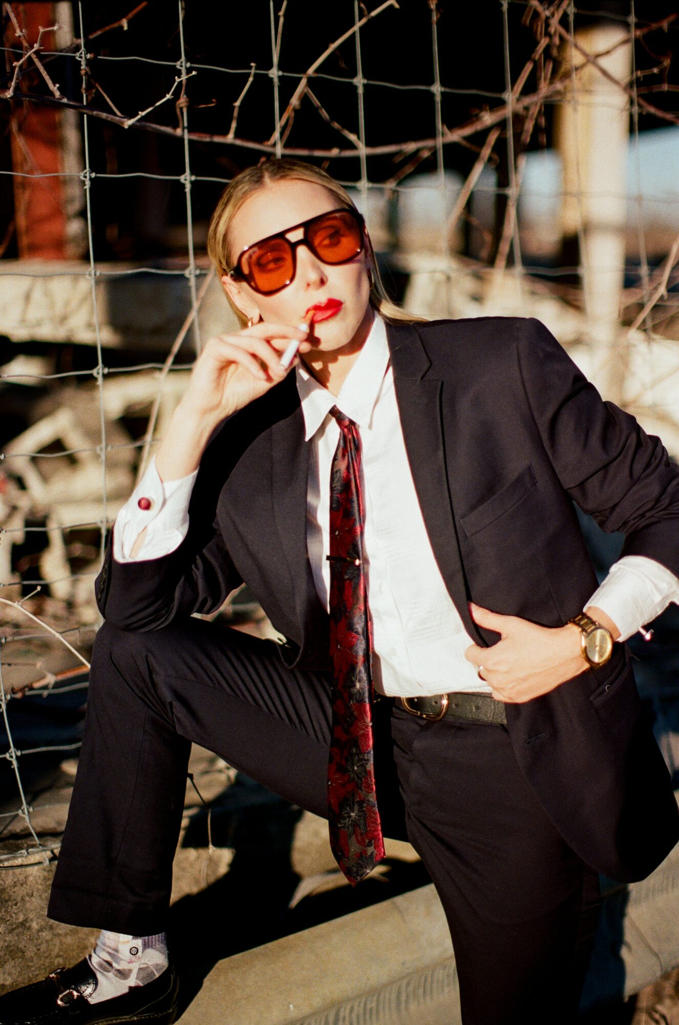 Woman in black suit and sunglasses sitting on a ledge in front of a net, with a city background.