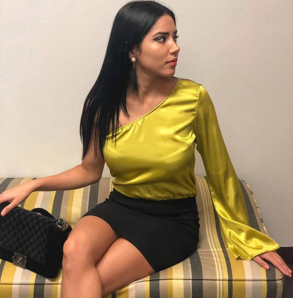 Woman with long black hair wearing a shiny yellow top and black skirt, sitting on a striped bench, looking to the side.