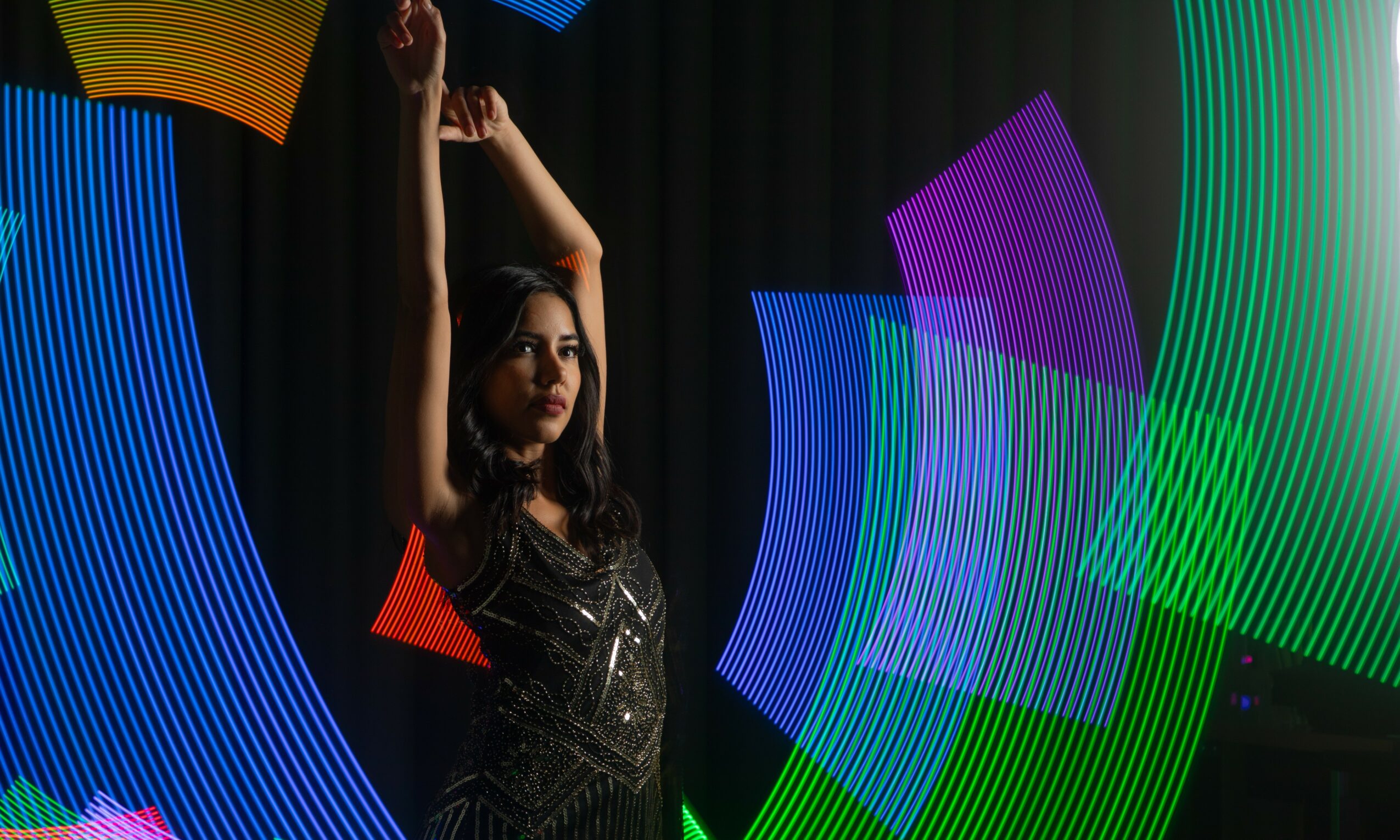 Woman with arms raised on stage surrounded by colorful light projections, dark background, wearing a black dress.