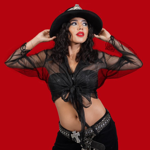 Woman with curly hair wearing a black hat, sheer black top, and black pants against a red background.