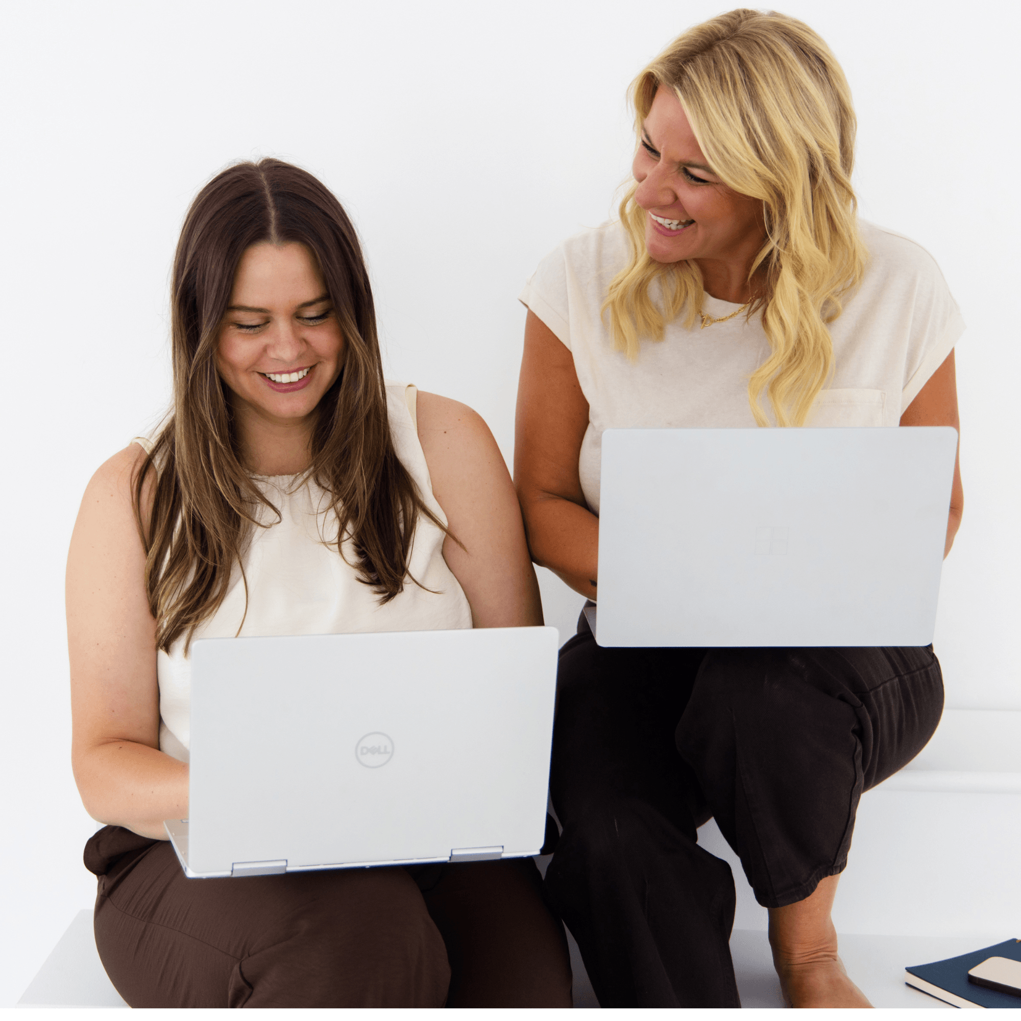 Two women sitting and smiling while using laptops, one with long dark hair and the other with blonde hair, against a white background.