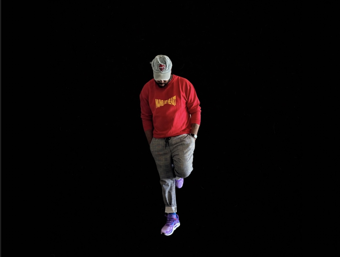 Person wearing a red sweatshirt, gray pants, purple shoes, and a white cap, standing against a black background.