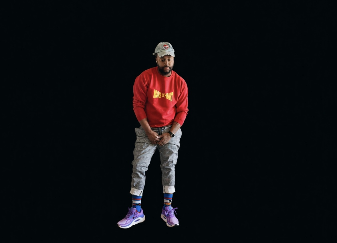 Person standing against a black background, wearing a red sweatshirt, gray pants, purple shoes, and a patterned cap.