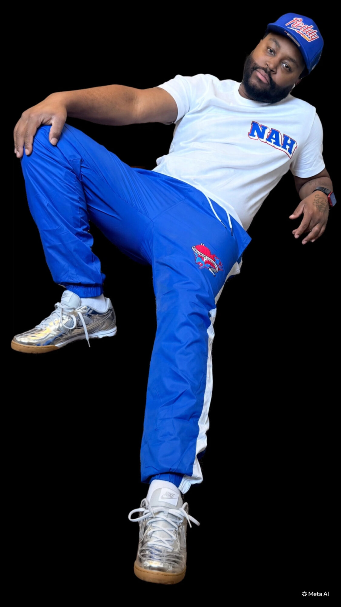 Person with a beard and cap, wearing a white T-shirt and blue pants, sitting with one leg raised, against a black background.