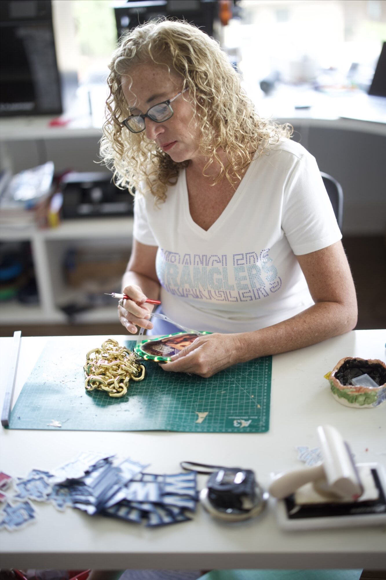 Woman with curly blonde hair and glasses working at a desk with a green cutting mat, handling a small object.