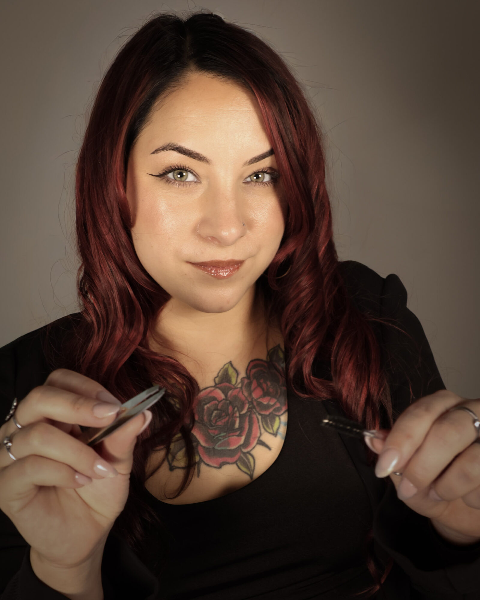 Woman with red hair and a tattoo on her chest, holding a pen and a small object, smiling at the camera.