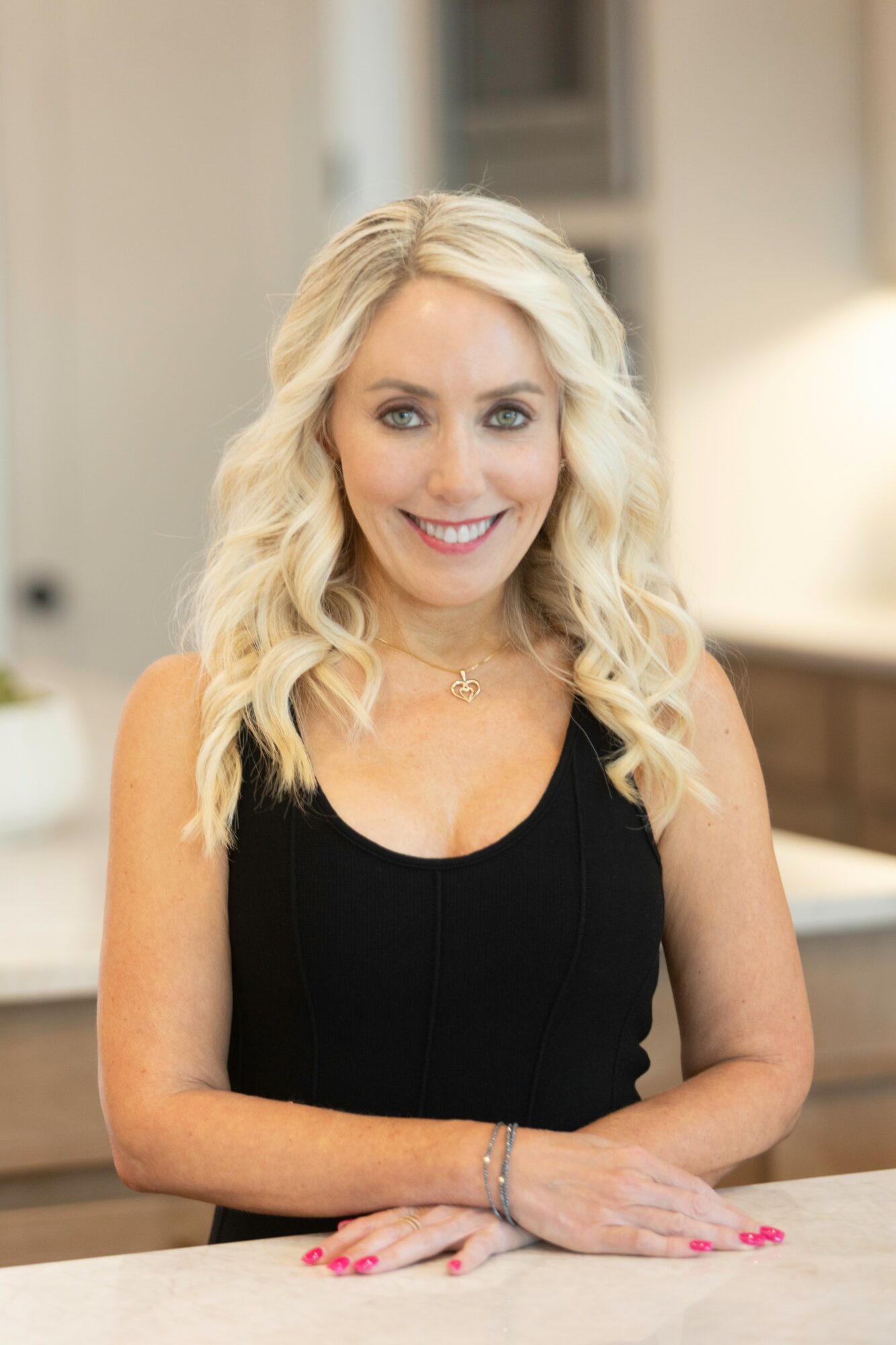 Smiling woman with blonde wavy hair wearing a black sleeveless top, sitting at a table indoors.