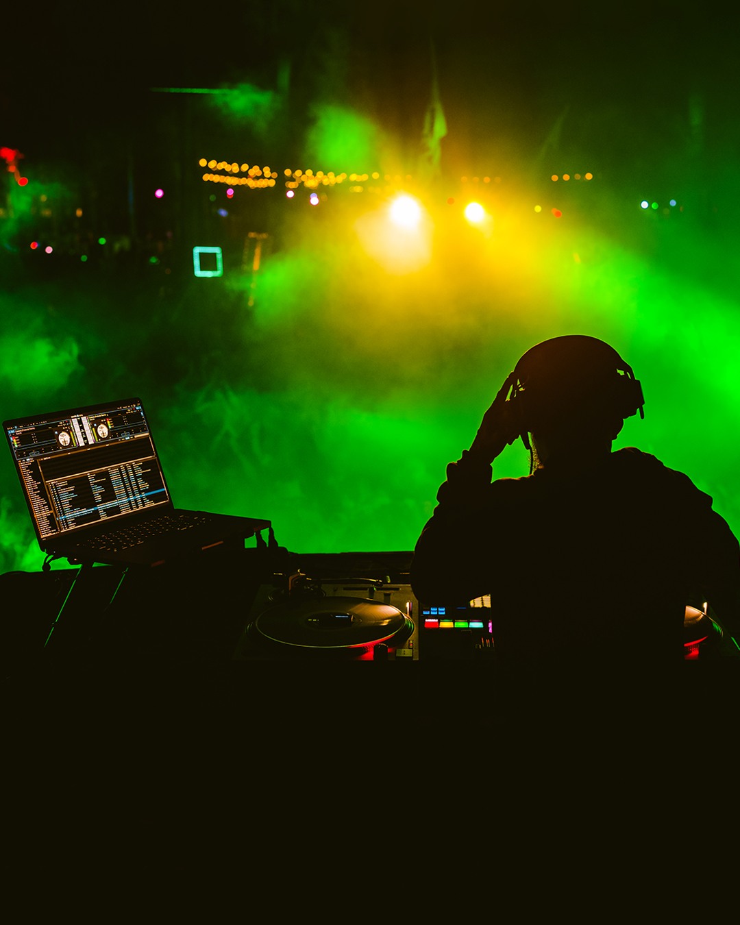DJ silhouette with equipment in front of colorful stage lights and fog at a concert or event.