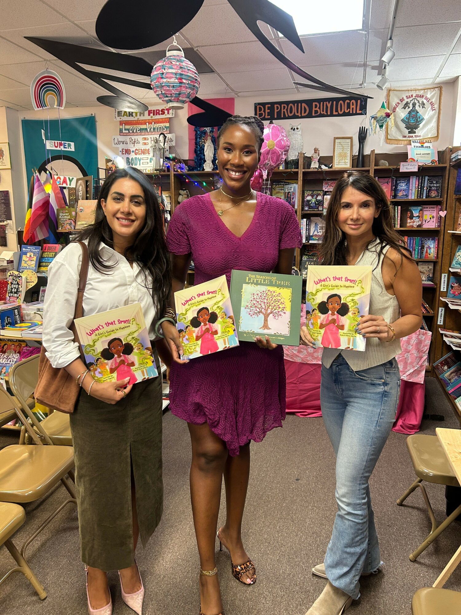 Three women holding children's books in a bookstore, smiling, with bookshelves and decorations in the background.