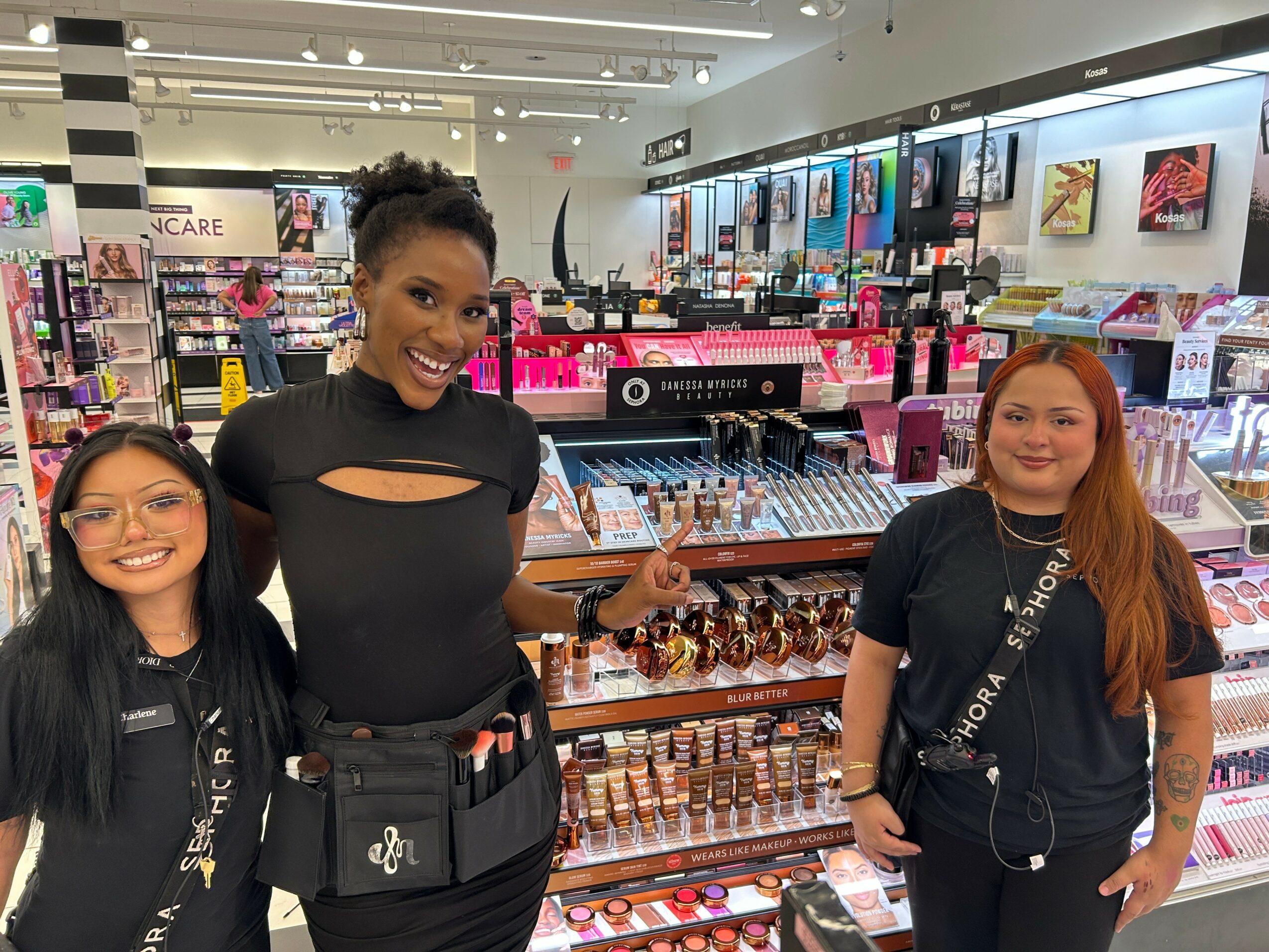Three women standing in a cosmetics store with shelves of makeup and beauty products behind them.