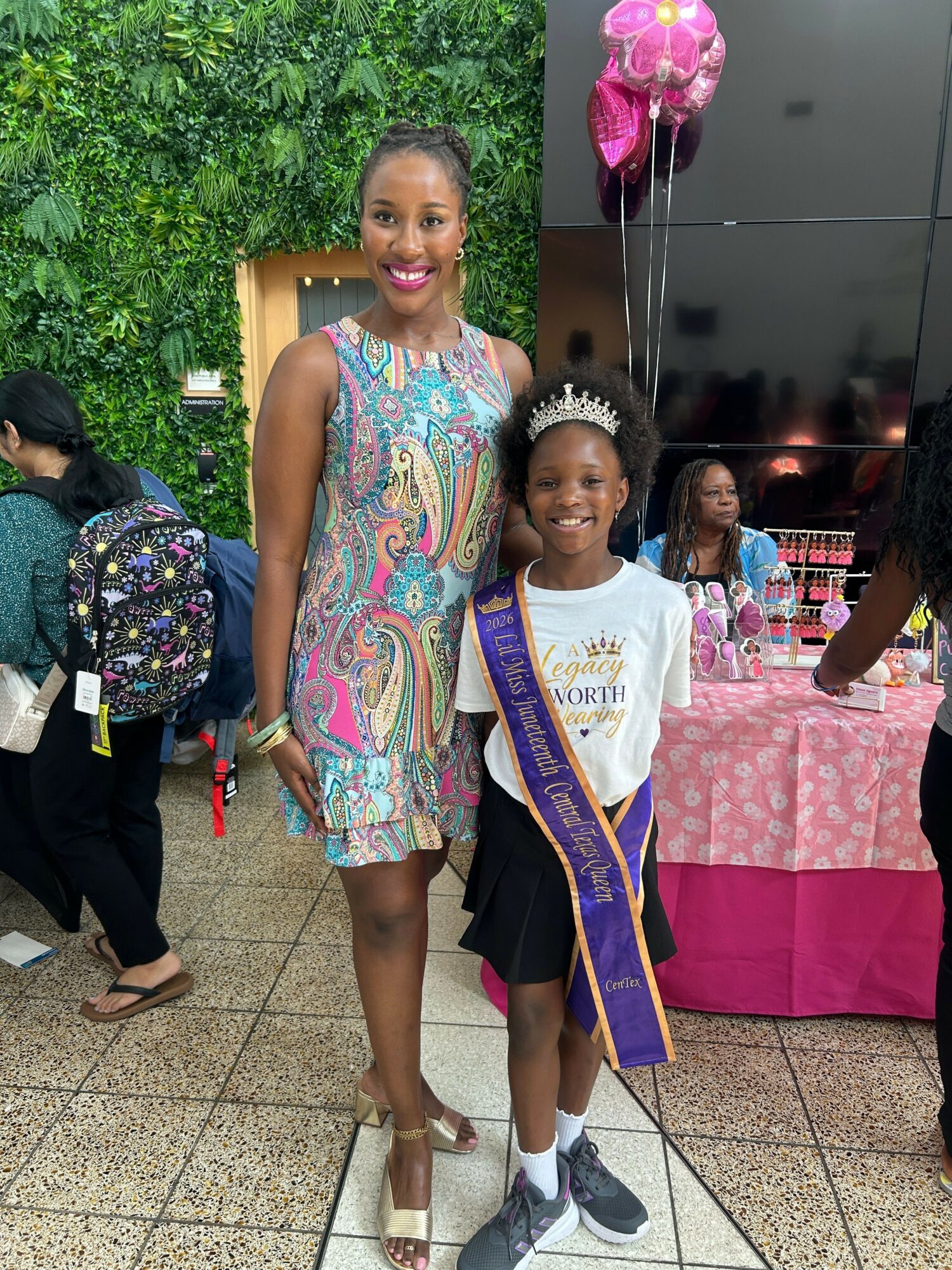 Girl in a white shirt and sash standing next to a woman in a colorful dress, with a decorated background and balloons.
