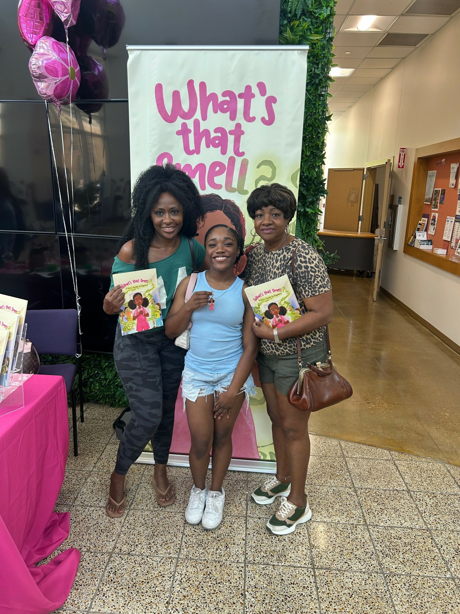 Three women standing outdoors, holding colorful gift bags, smiling, with a sign and pink balloons in the background.