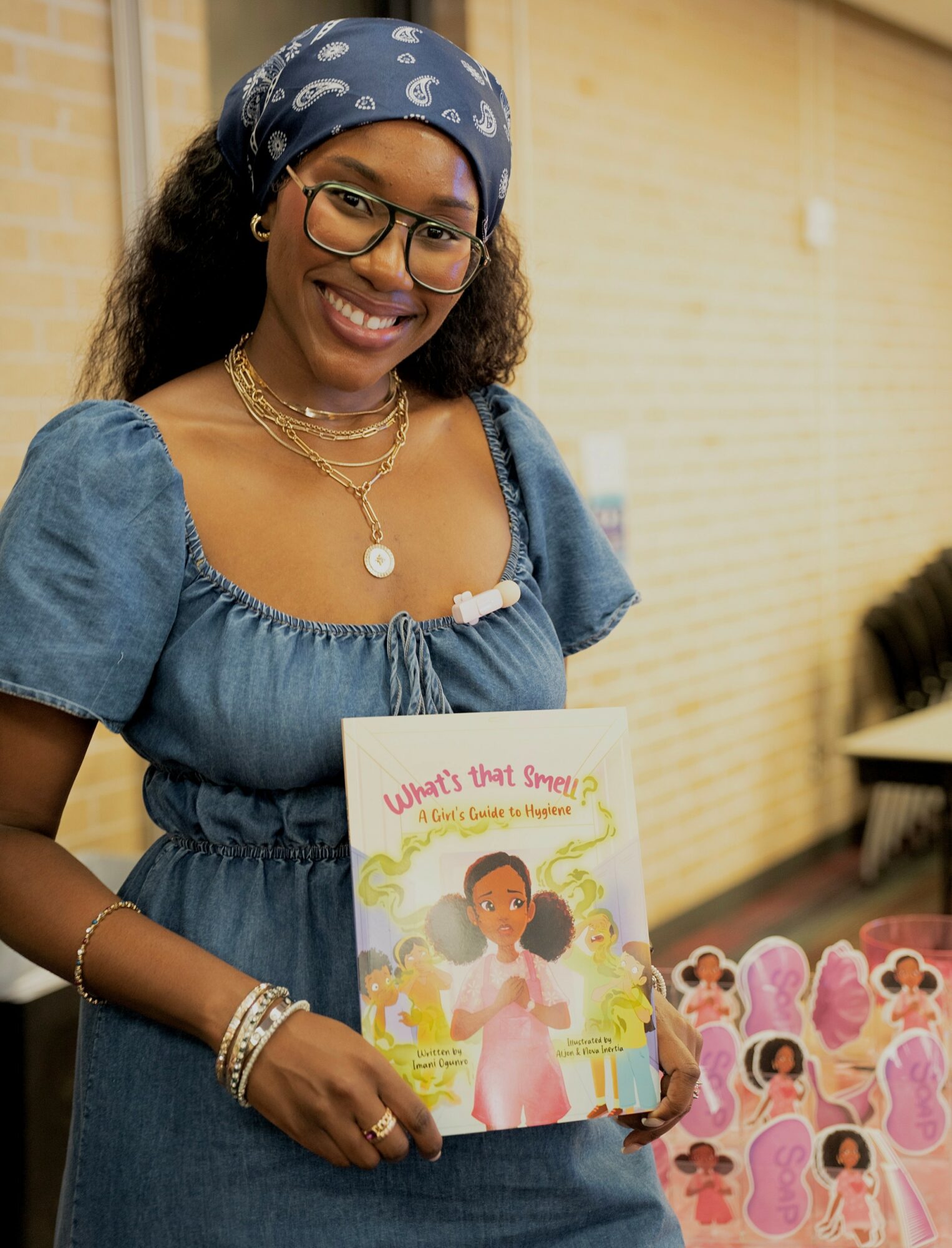 Smiling woman with glasses and a blue headscarf holding a children's book, wearing a blue dress and jewelry, in a room with a yellow wall.