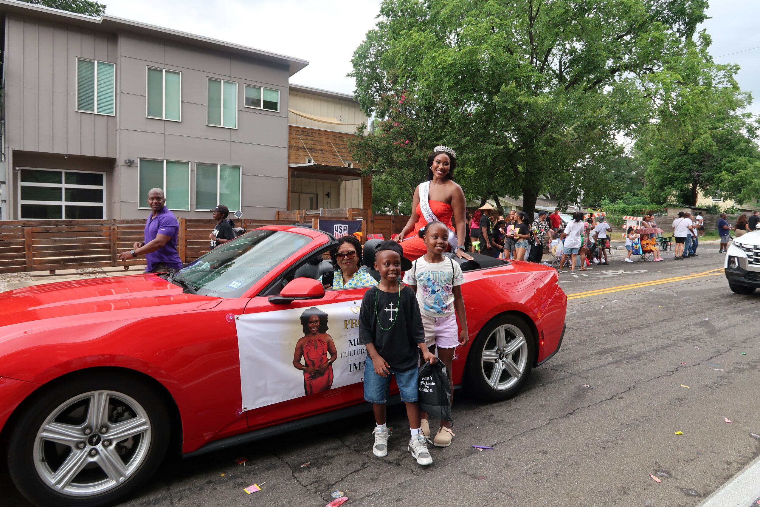 Group of children and a woman standing next to a red convertible car on a street with people and trees in the background.