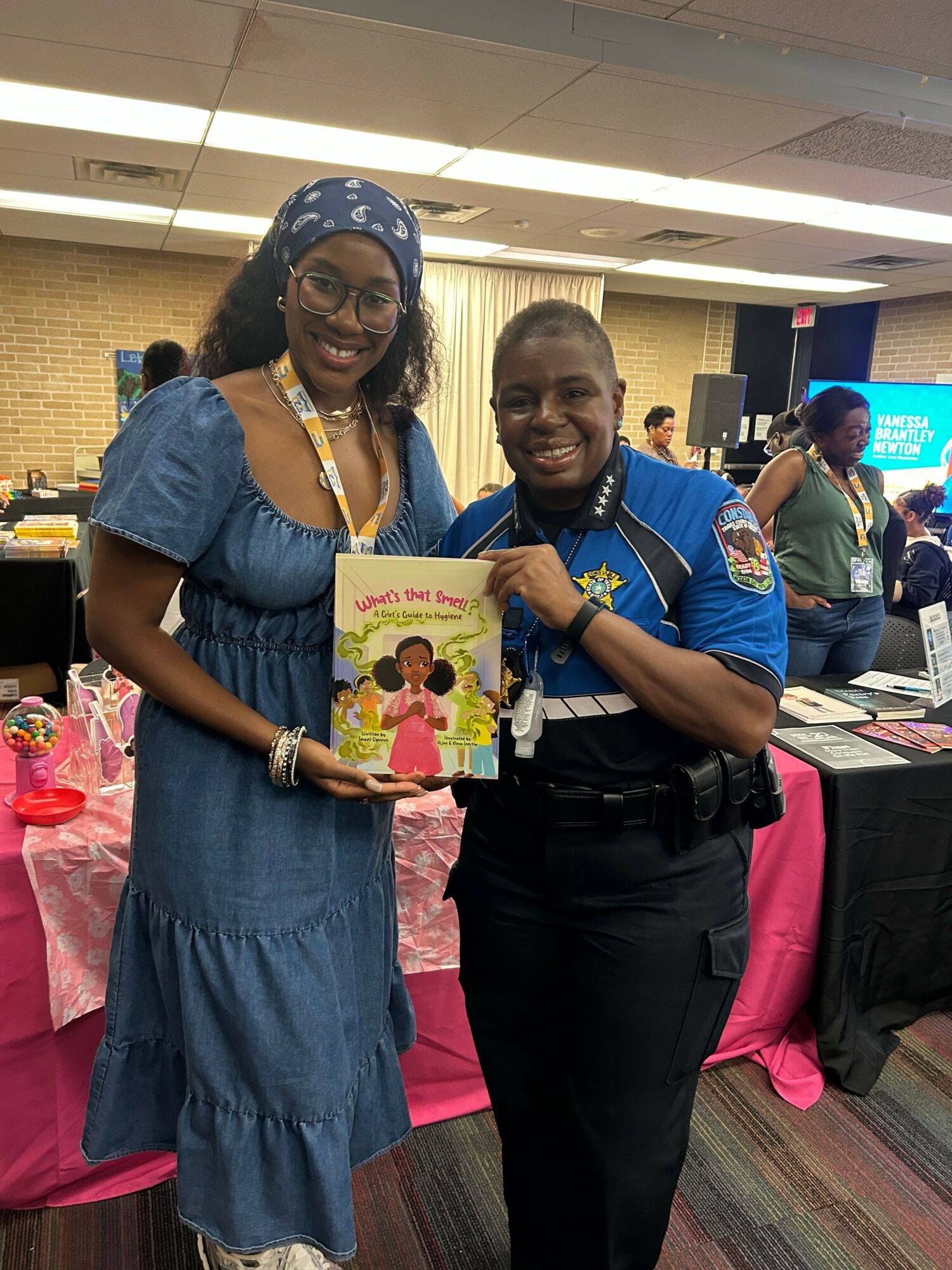 Two women standing indoors, one holding a colorful children's book, smiling at the camera, with tables and people in the background.