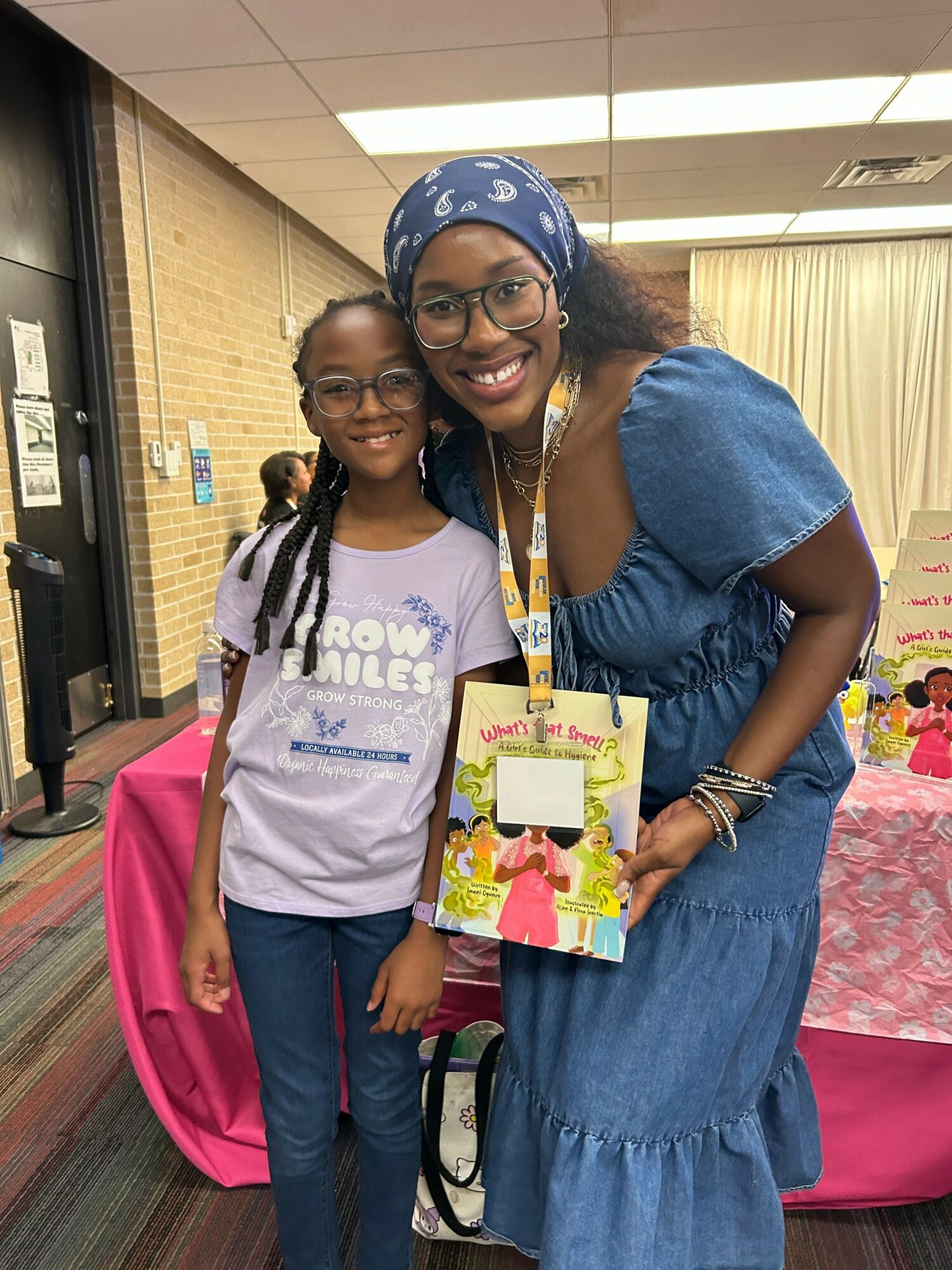 Two women smiling, one wearing glasses and a headscarf, the other with braids, standing indoors with a pink tablecloth and flowers behind them.