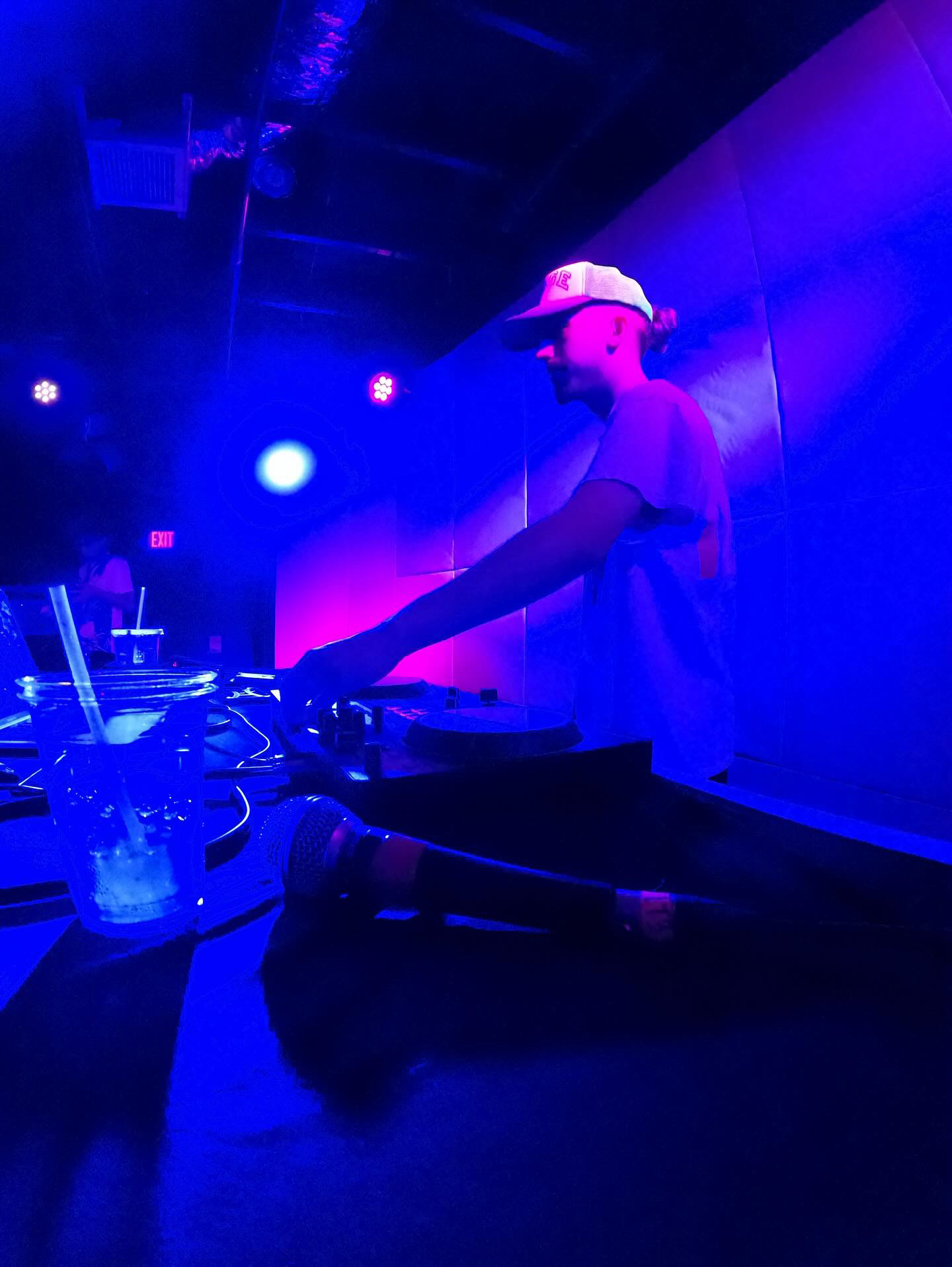DJ wearing a cap performs at a nightclub with blue and purple lighting, equipment on a table, and a dark background.