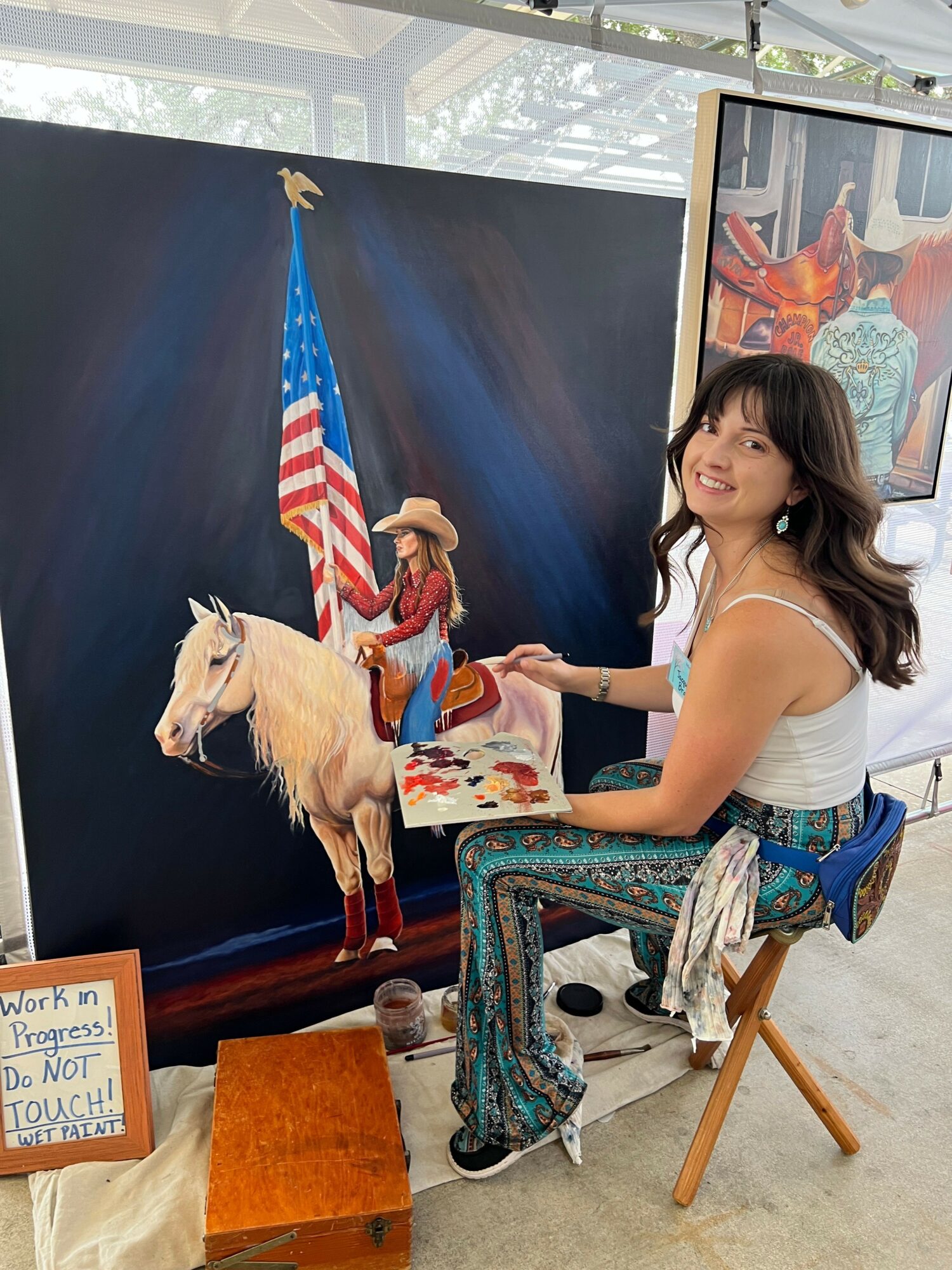 Woman with long dark hair in a white top and patterned pants stands next to a large painting of a dog with an American flag hat, holding a paintbrush.