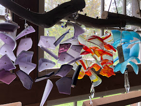 Colorful wind chimes hanging indoors near a window with trees outside, made of various shaped plastic pieces in purple, white, red, orange, and blue.