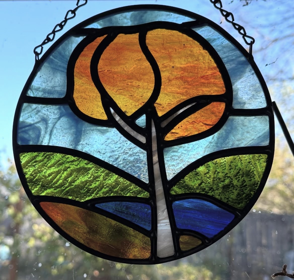 Stained glass circular window with a tree, sun, green hills, and water scene, hanging outdoors with sky background.