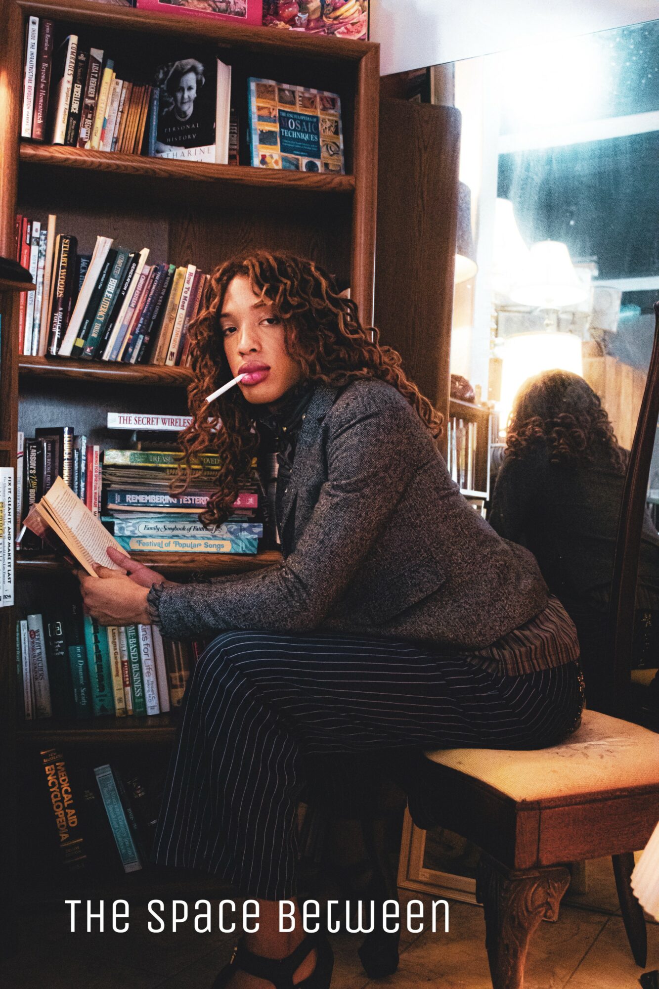 Woman with curly hair reading a book in a library, sitting on a wooden chair, bookshelf behind her, sunlight through window.