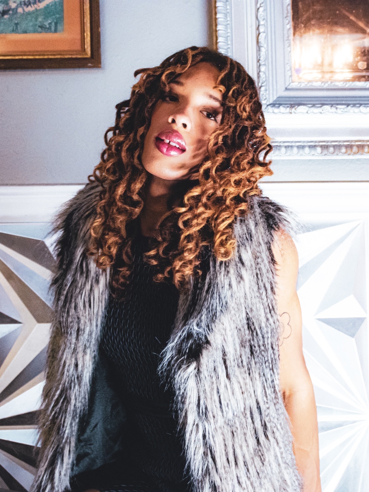Woman with curly hair wearing a fur vest, standing indoors with decorative wall panels and framed artwork behind her.