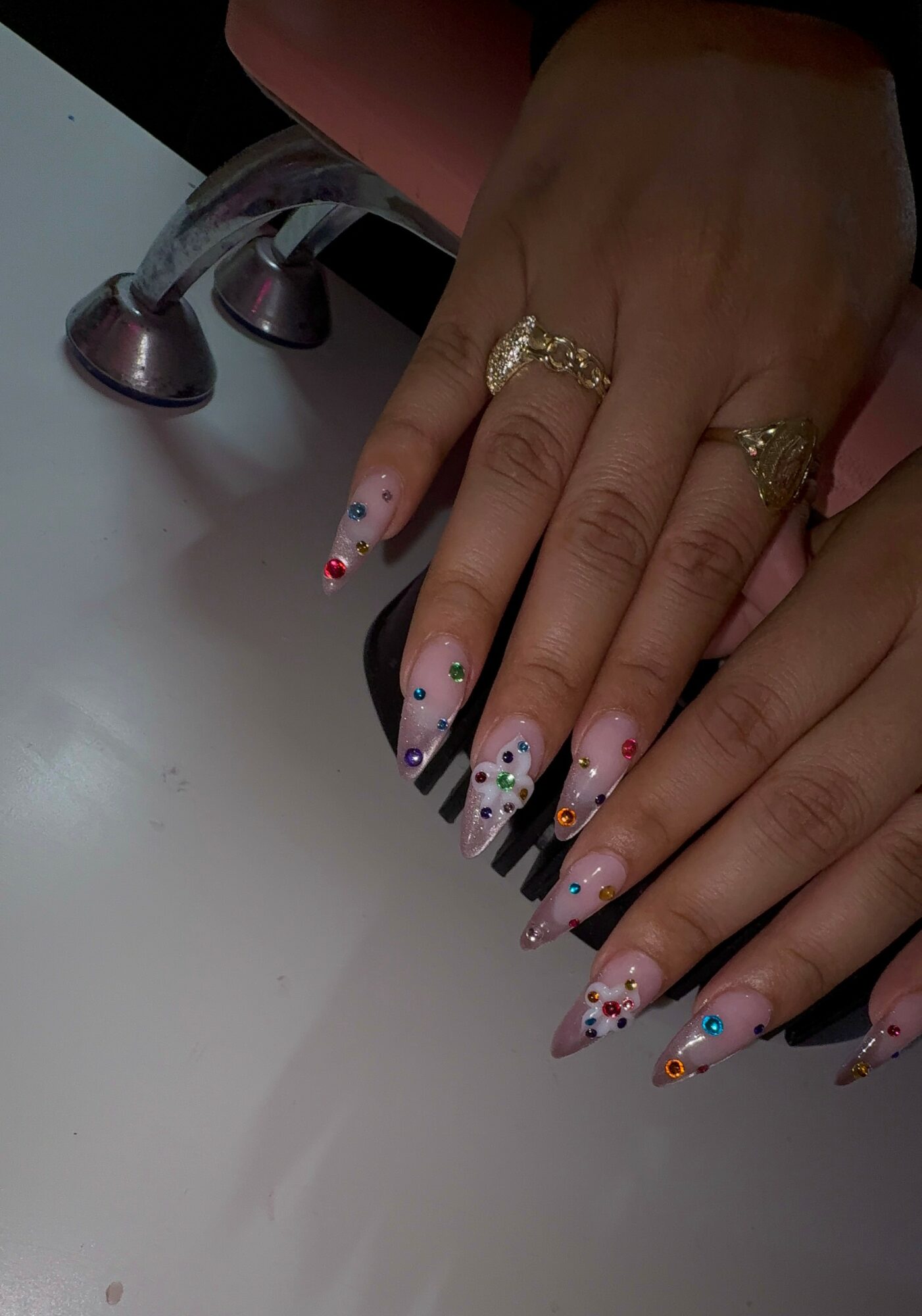Hands with decorated nails resting on a surface, wearing multiple rings, with a faucet in the background.