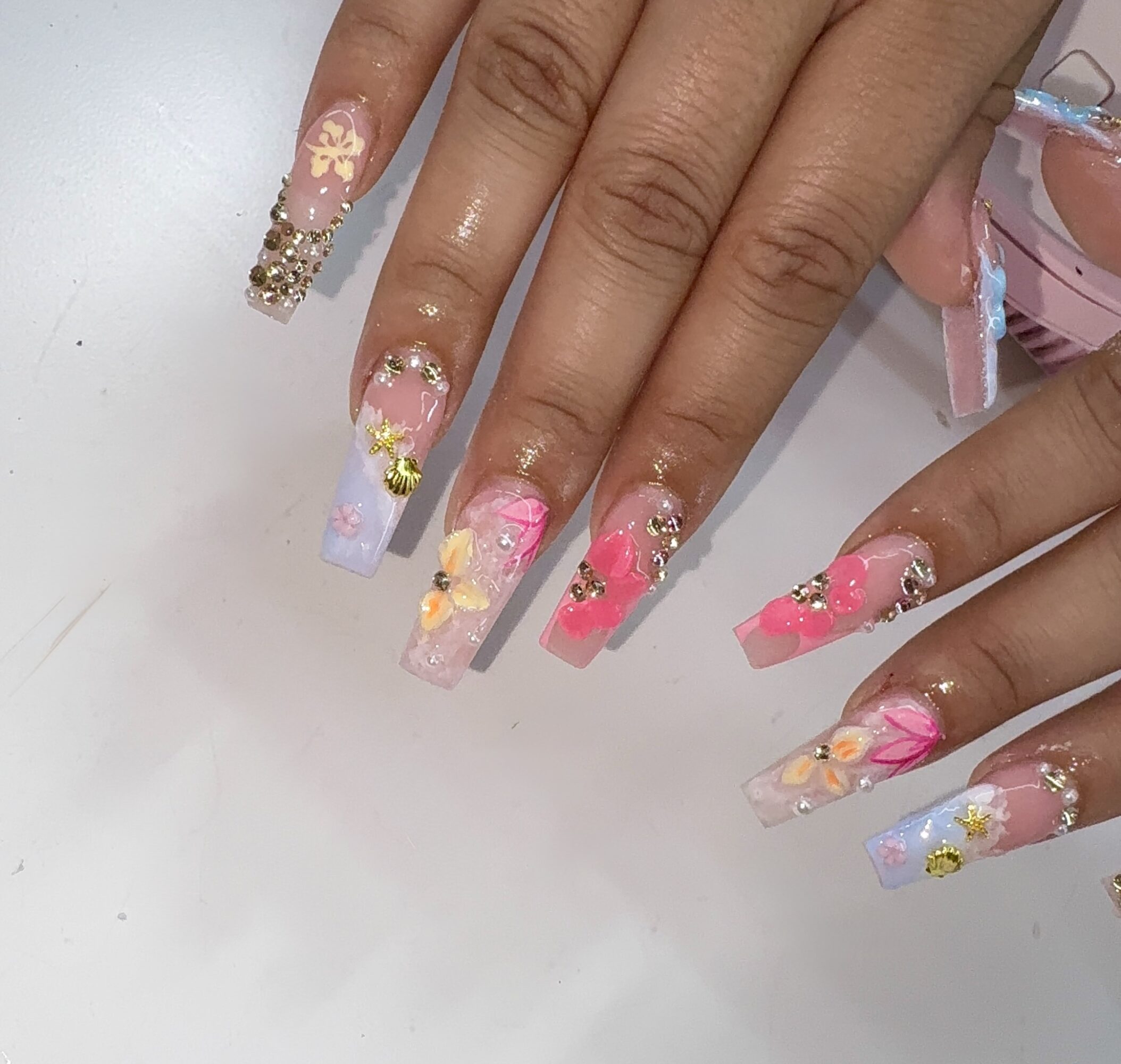 Hand with decorated long nails featuring pink, blue, and clear designs with floral and gold accents, on a white surface.