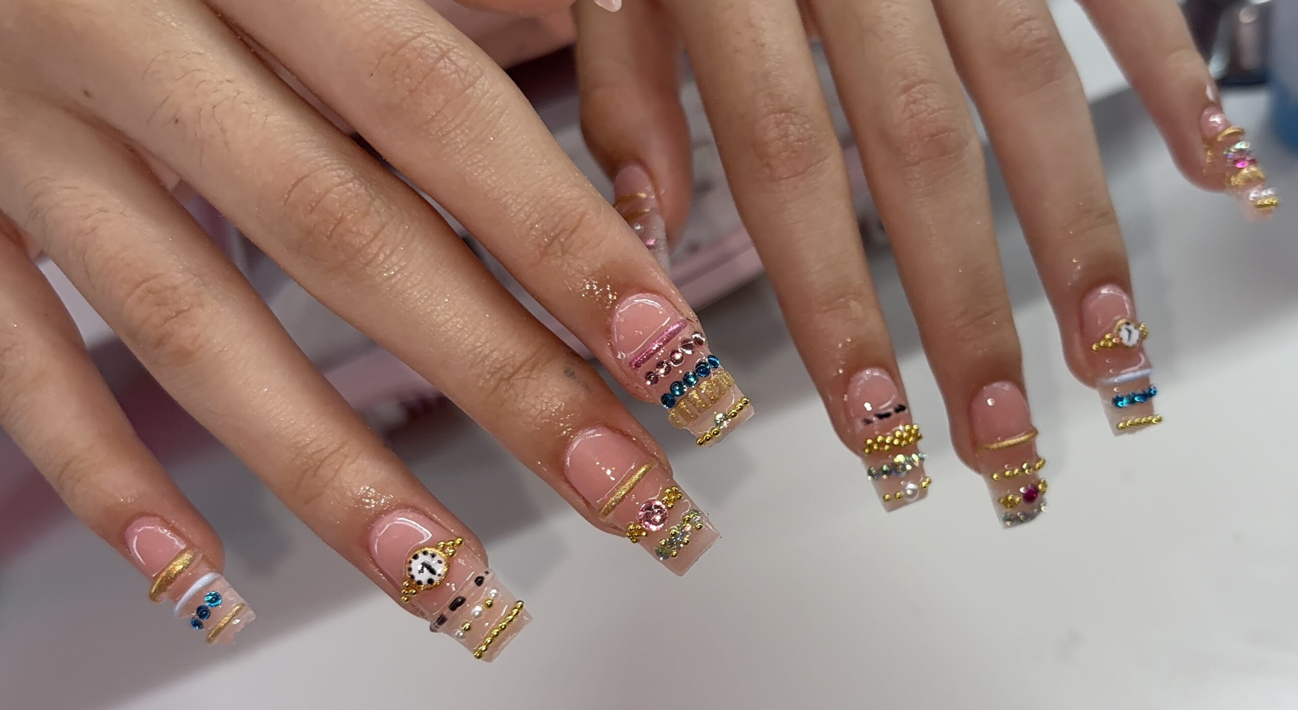 Close-up of hands with decorated nails featuring pink, gold, and blue designs, and rhinestones, on a white surface.