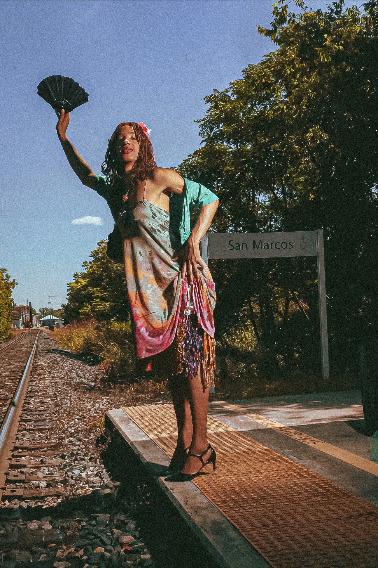 Woman standing on a platform near train tracks, holding a fan, with trees and a sign reading 'San Marcos' in the background.