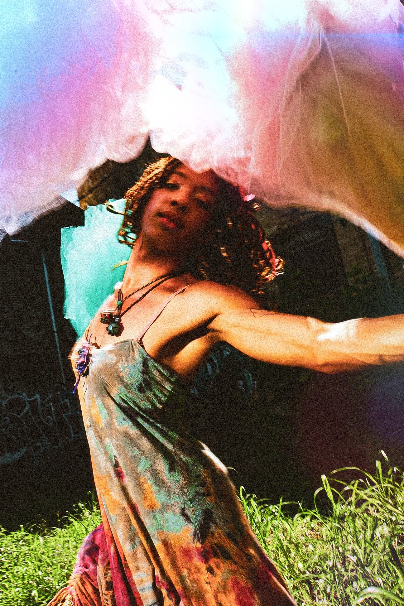 Woman with curly hair in a colorful dress outdoors, arms outstretched, under a pink and yellow canopy, grass in foreground.