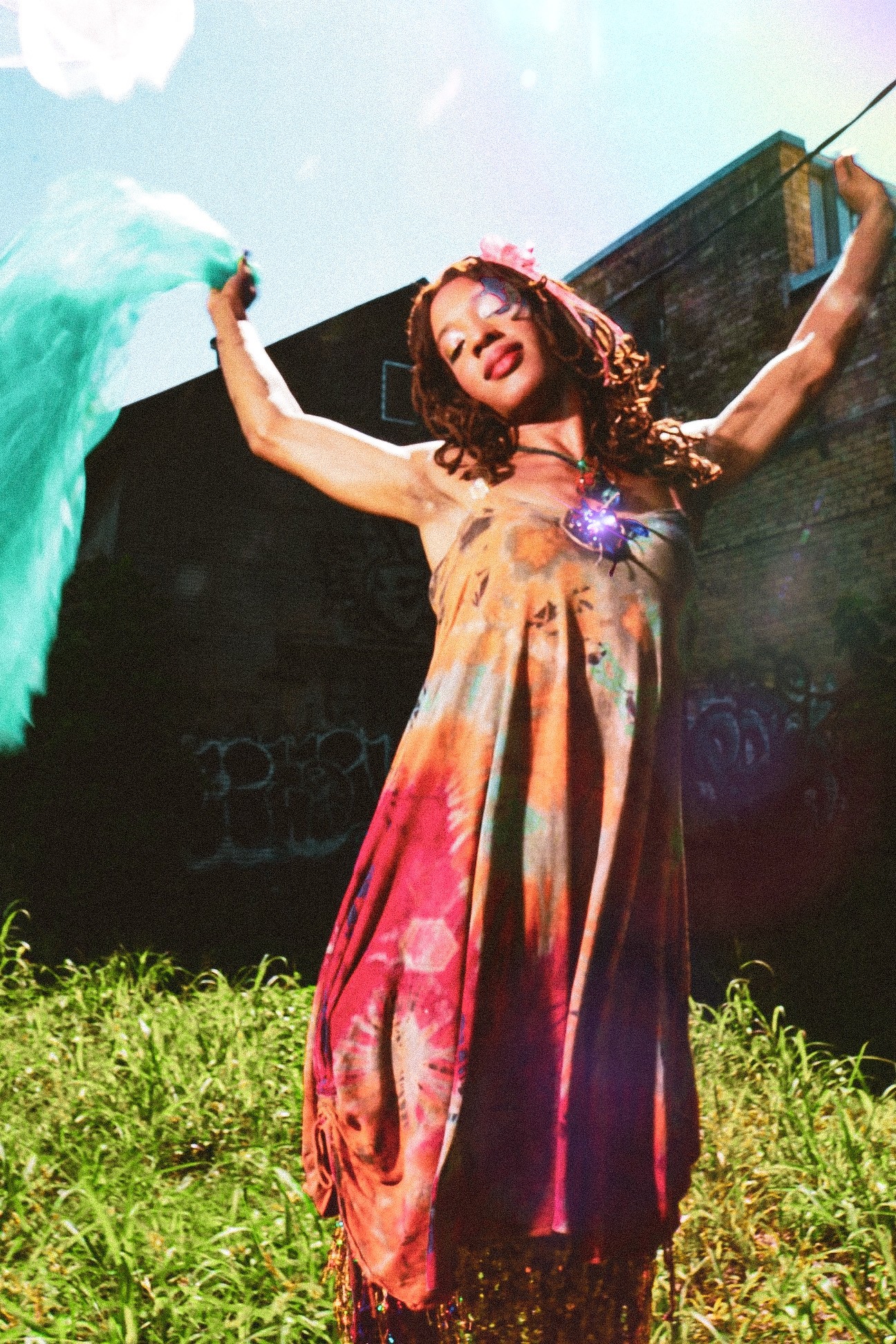 Woman in colorful dress with arms raised outdoors, holding fabric, smiling, with a building and blue sky in background.