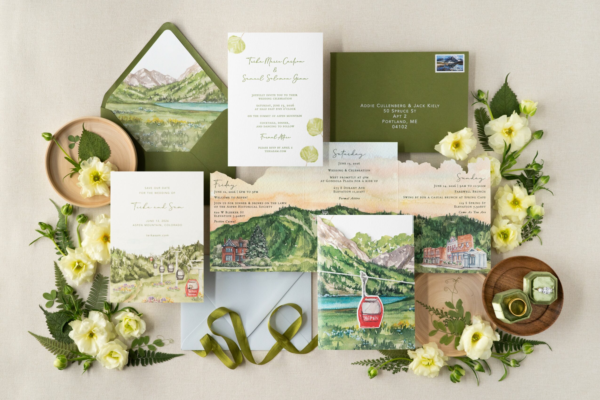 Wedding invitation suite with greenery and white flowers, including cards, envelopes, and decorative elements, arranged on a light background.