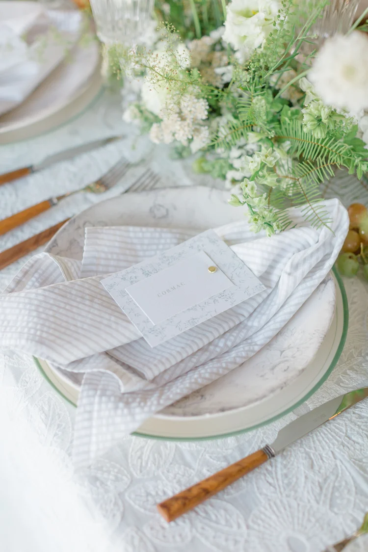 A white cloth napkin with a tag, placed on a plate with a floral arrangement nearby, on a table setting.