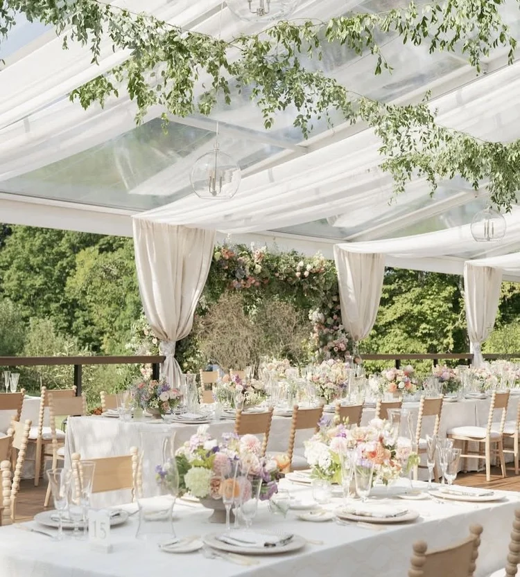 Elegant outdoor event setup with decorated tables, floral arrangements, and draped canopy, surrounded by greenery.
