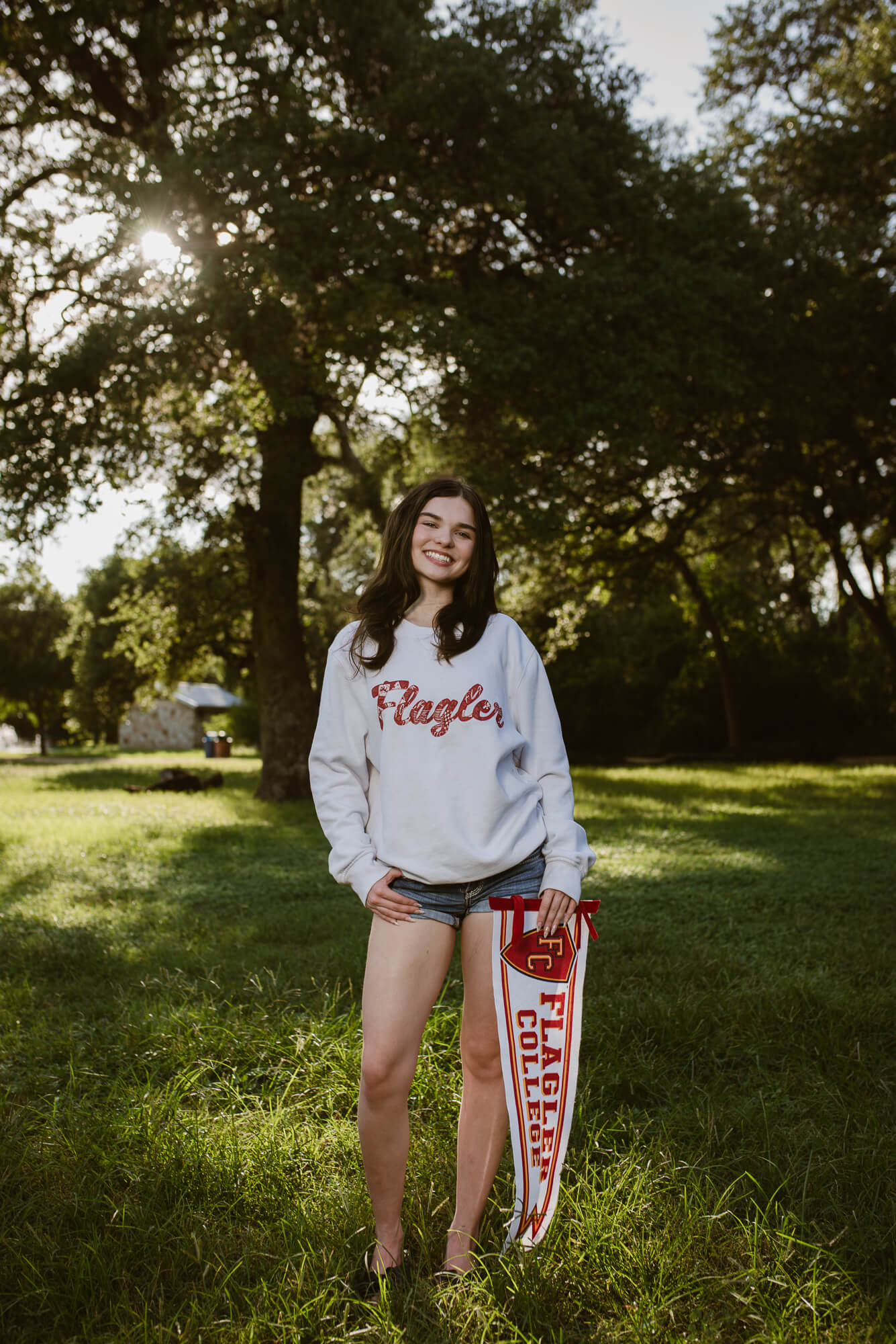 Young woman standing outdoors in a park with trees and sunlight, smiling, wearing a white sweatshirt and shorts, holding a skateboard.
