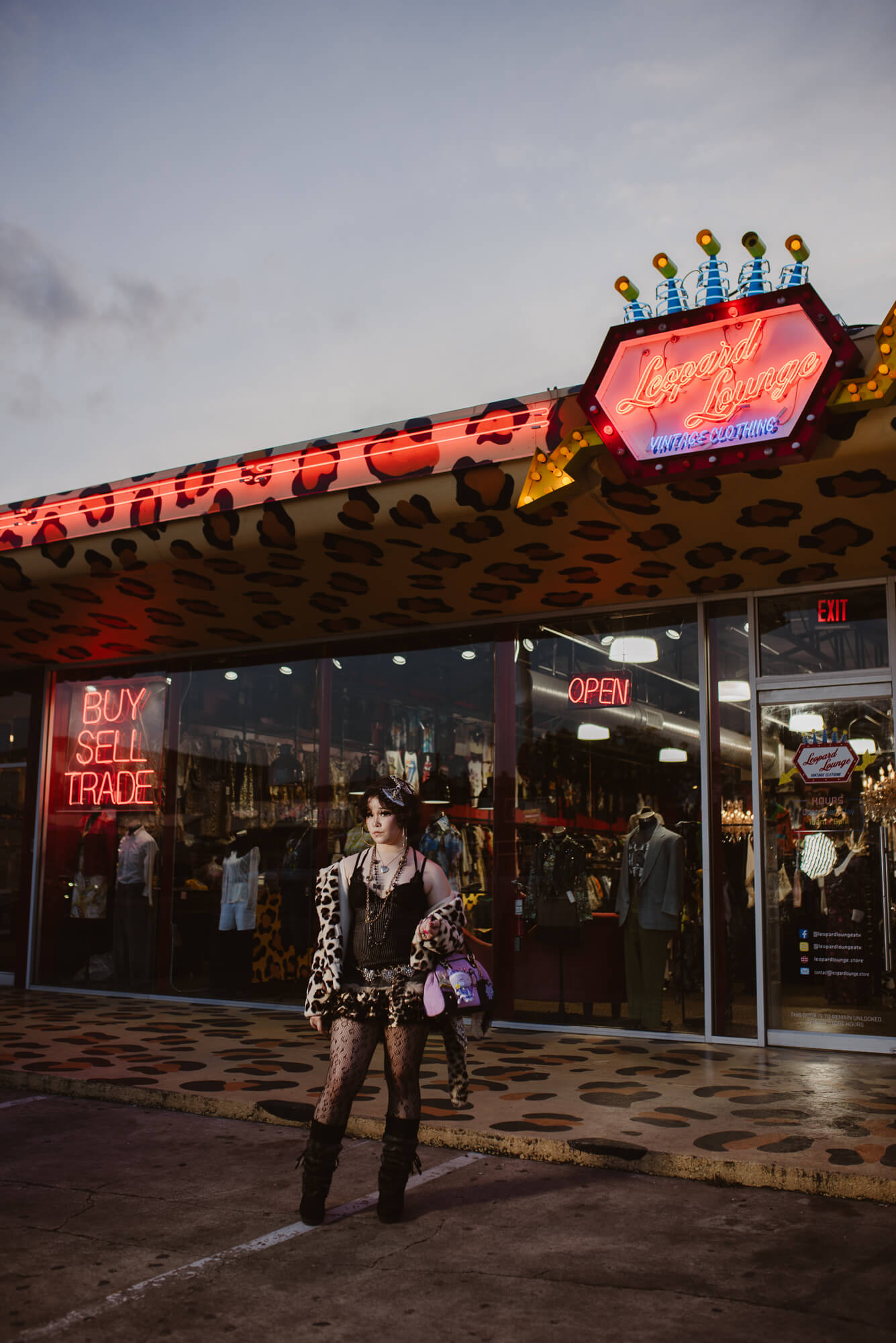 Person standing outside a store with neon signs and a leopard print canopy, holding a bag and umbrella, during evening or night.