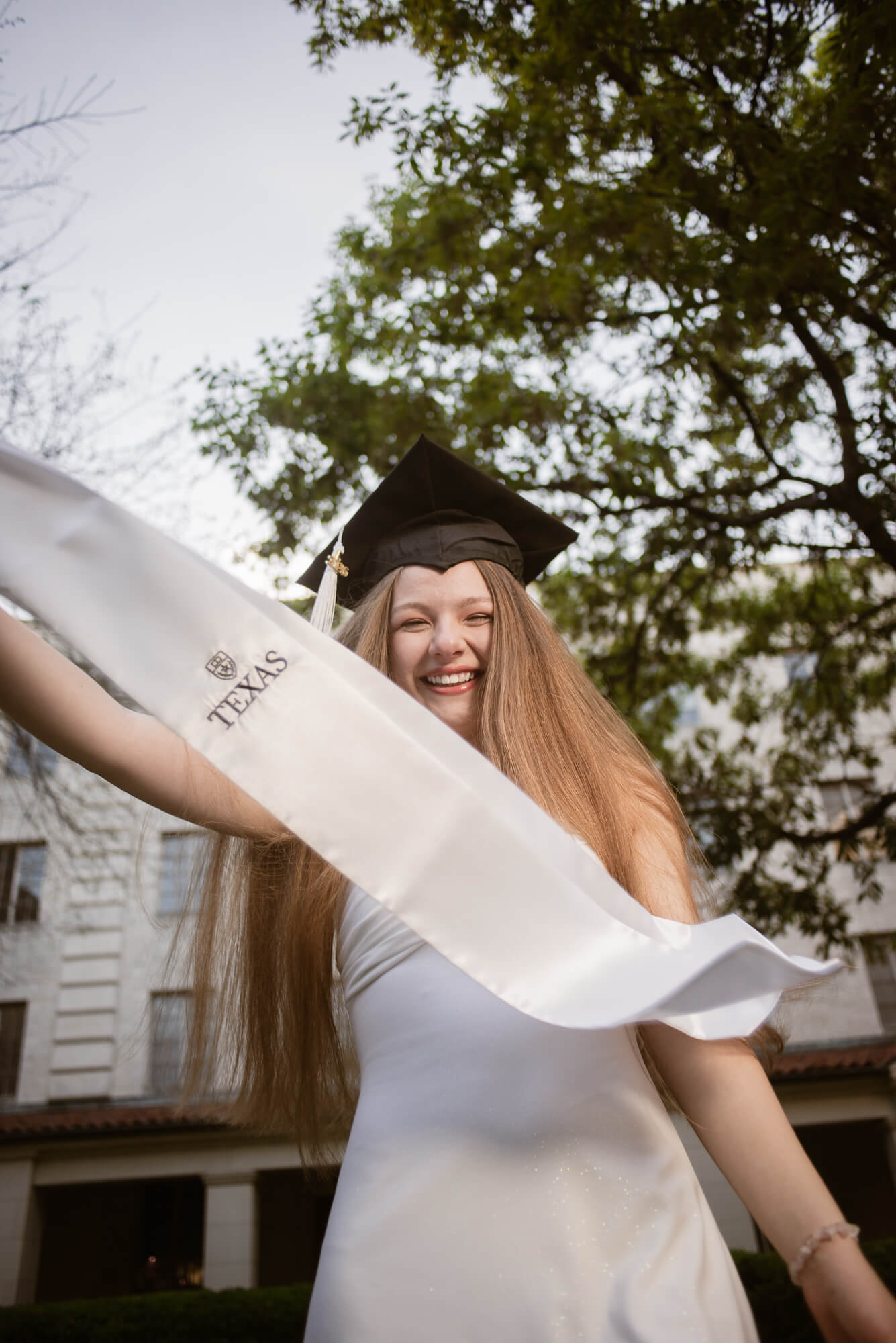 Young woman in graduation cap and gown smiling outdoors, holding a rolled-up diploma, trees and building in background.