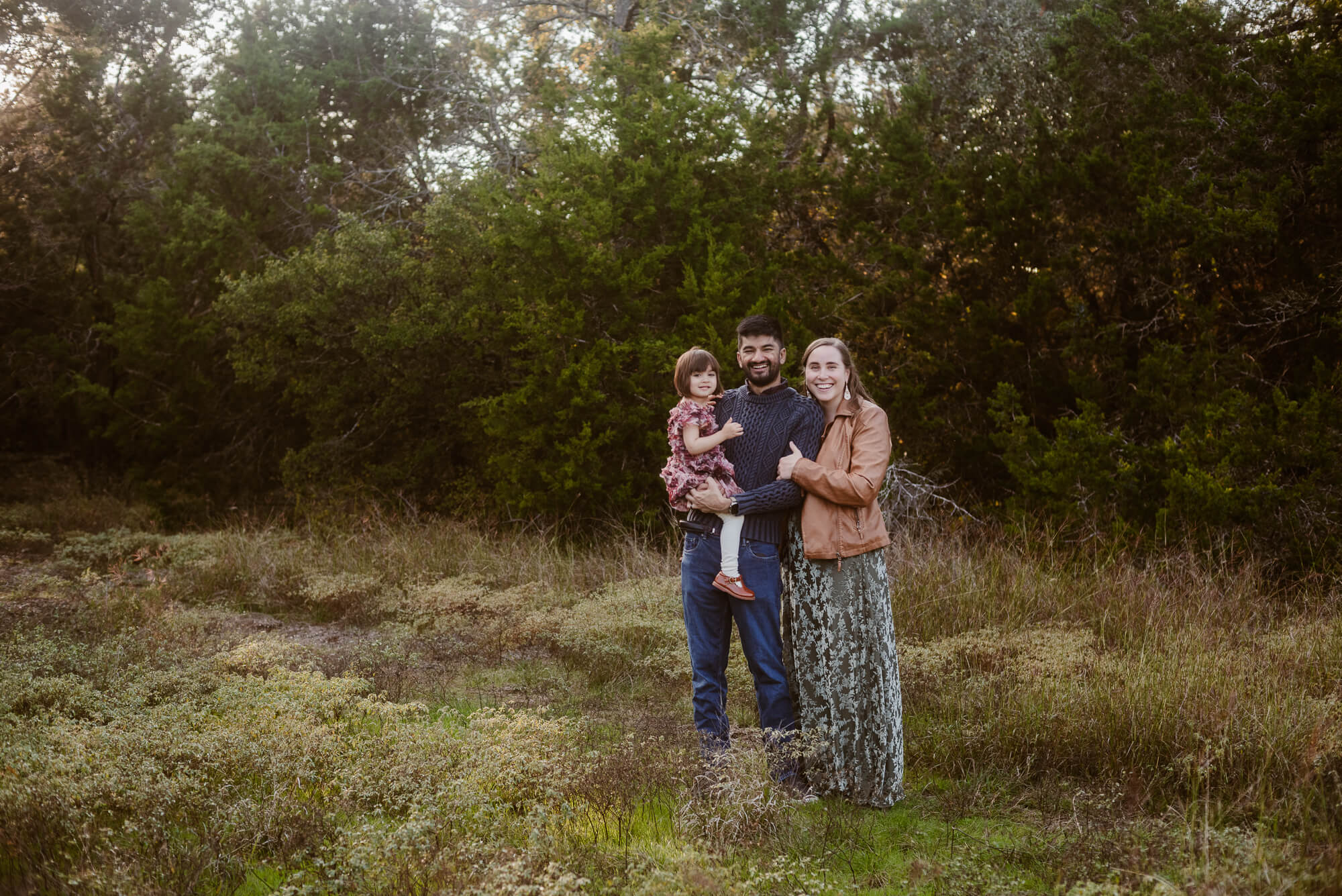Family of three standing outdoors in a grassy area with trees in the background, smiling at the camera.