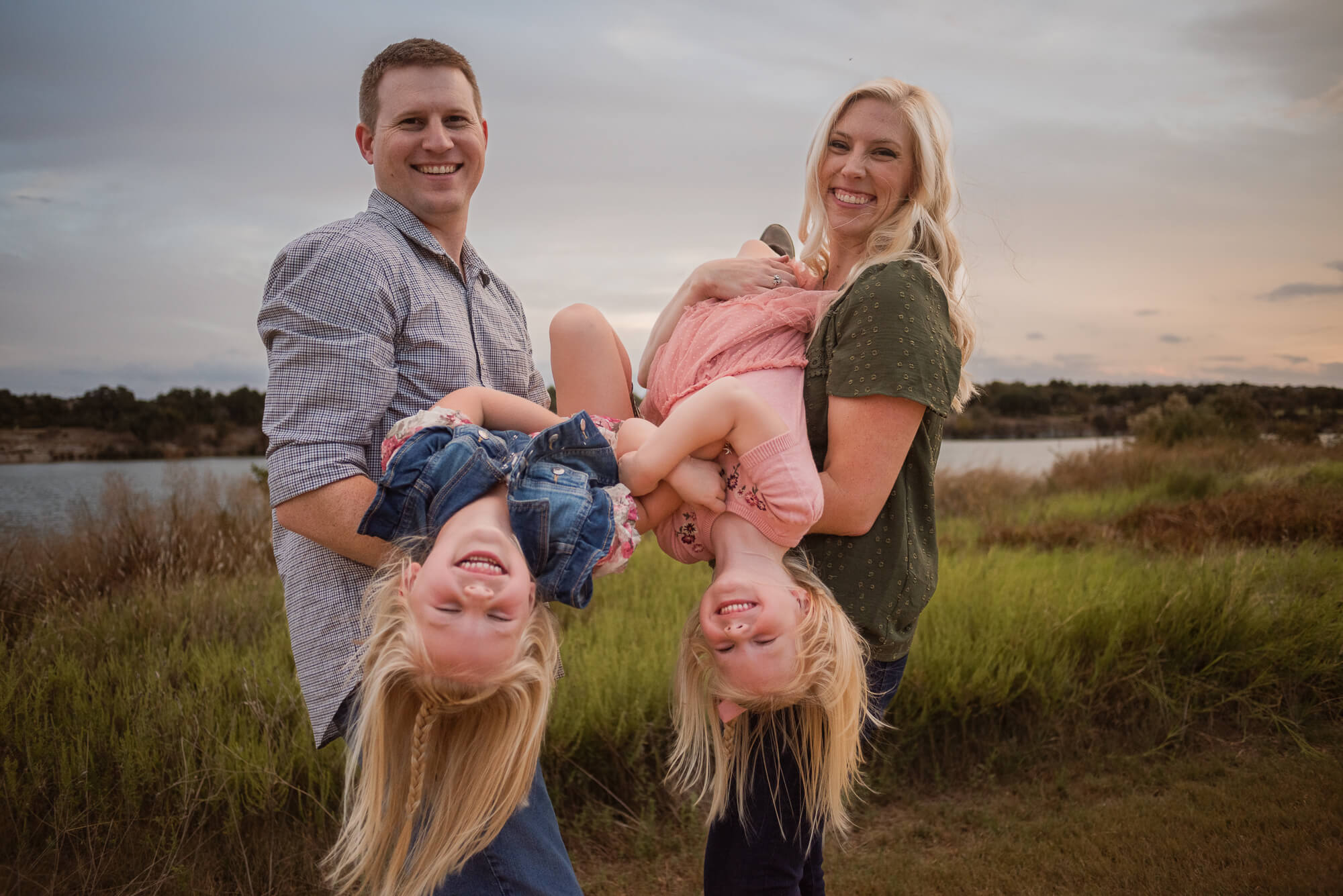 Four people outdoors, two adults holding two children upside down, smiling, with a grassy field and water in the background.