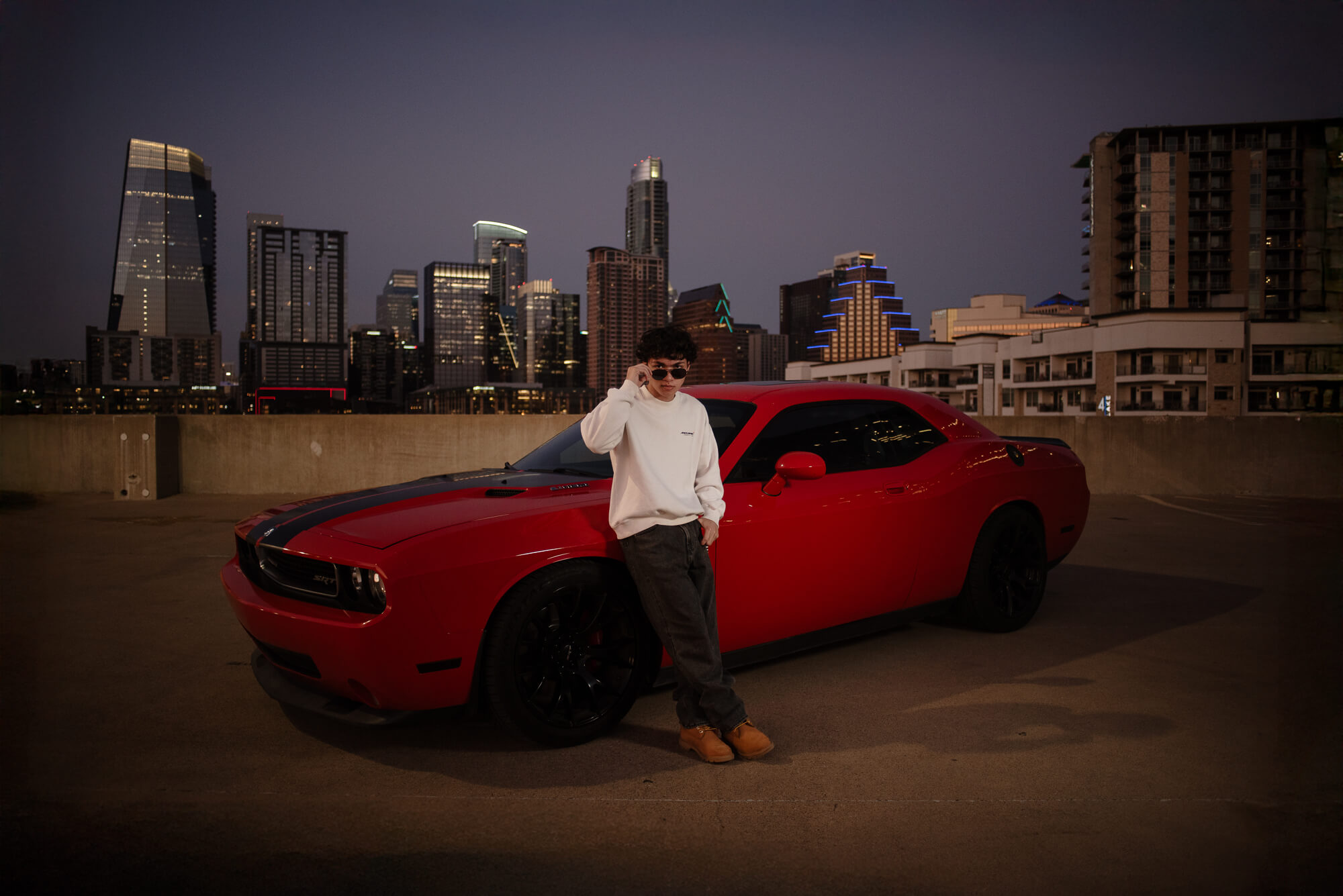 Person standing next to a red car on a rooftop parking lot with city skyline at night.